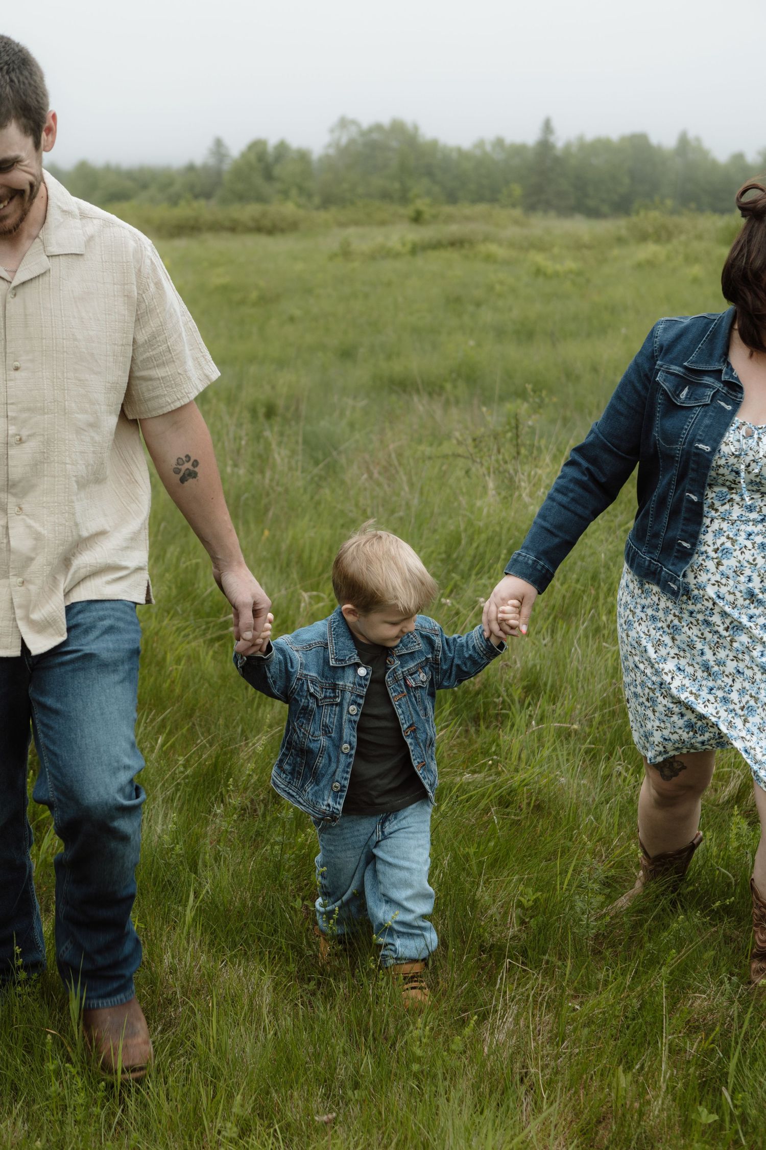 A family walks hand-in-hand through a lush green field, parents swinging their toddler in a denim jacket. New Brunswick Family Photography
