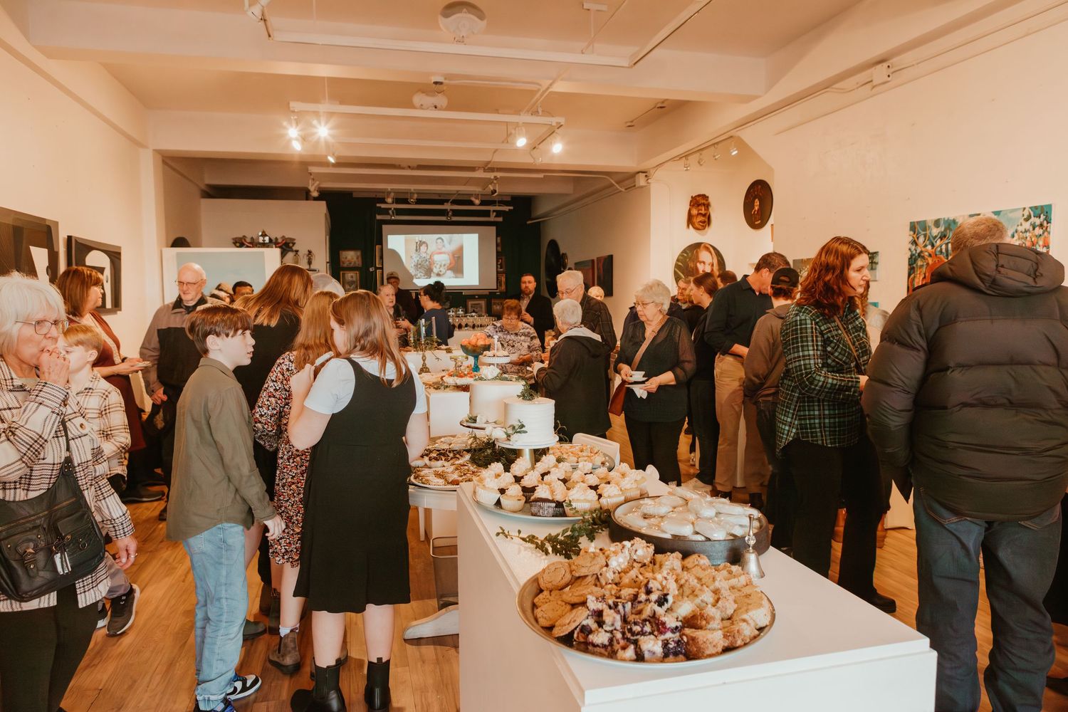 Guests mingle at a bustling art gallery, with food displayed on white tables and artwork hanging on the walls.