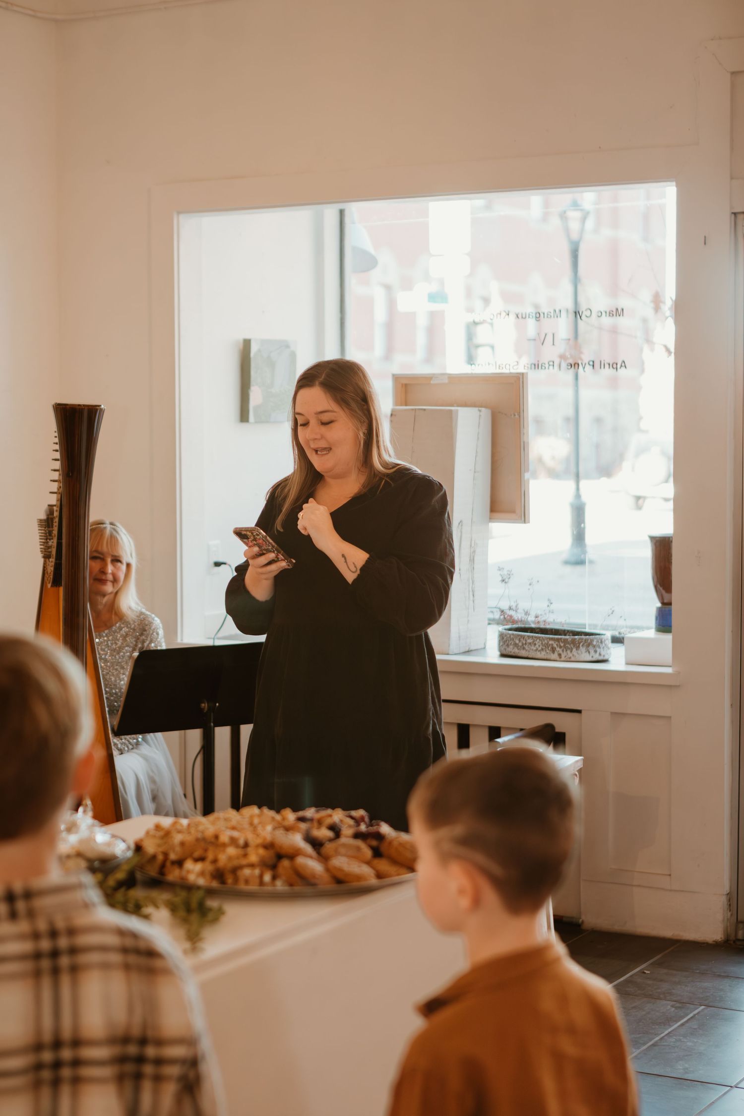 Woman in black dress reads from phone at indoor gathering with food display and children nearby.