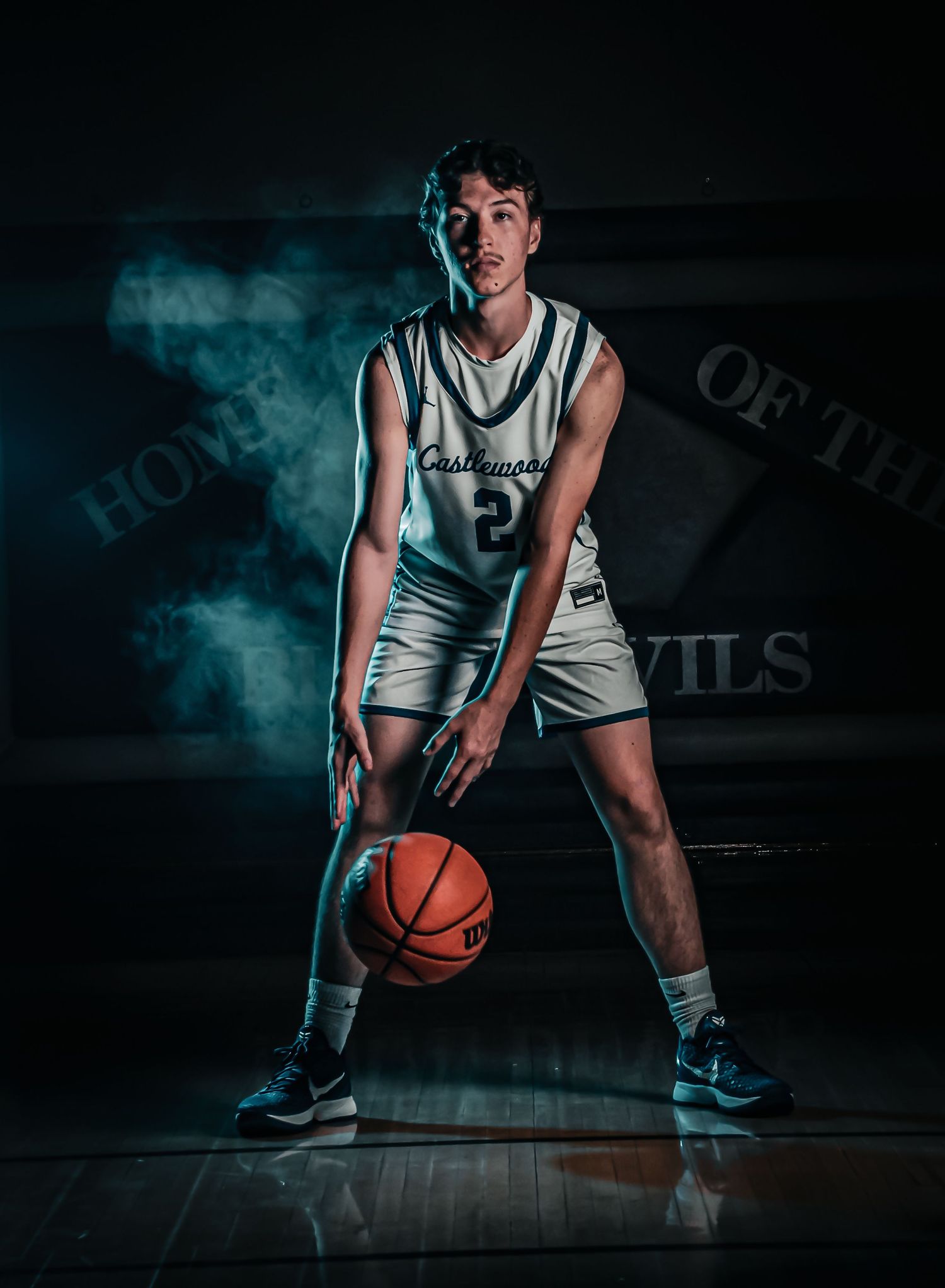 Basketball player dribbles in athletic stance on dark court with moody lighting and smoke effects.