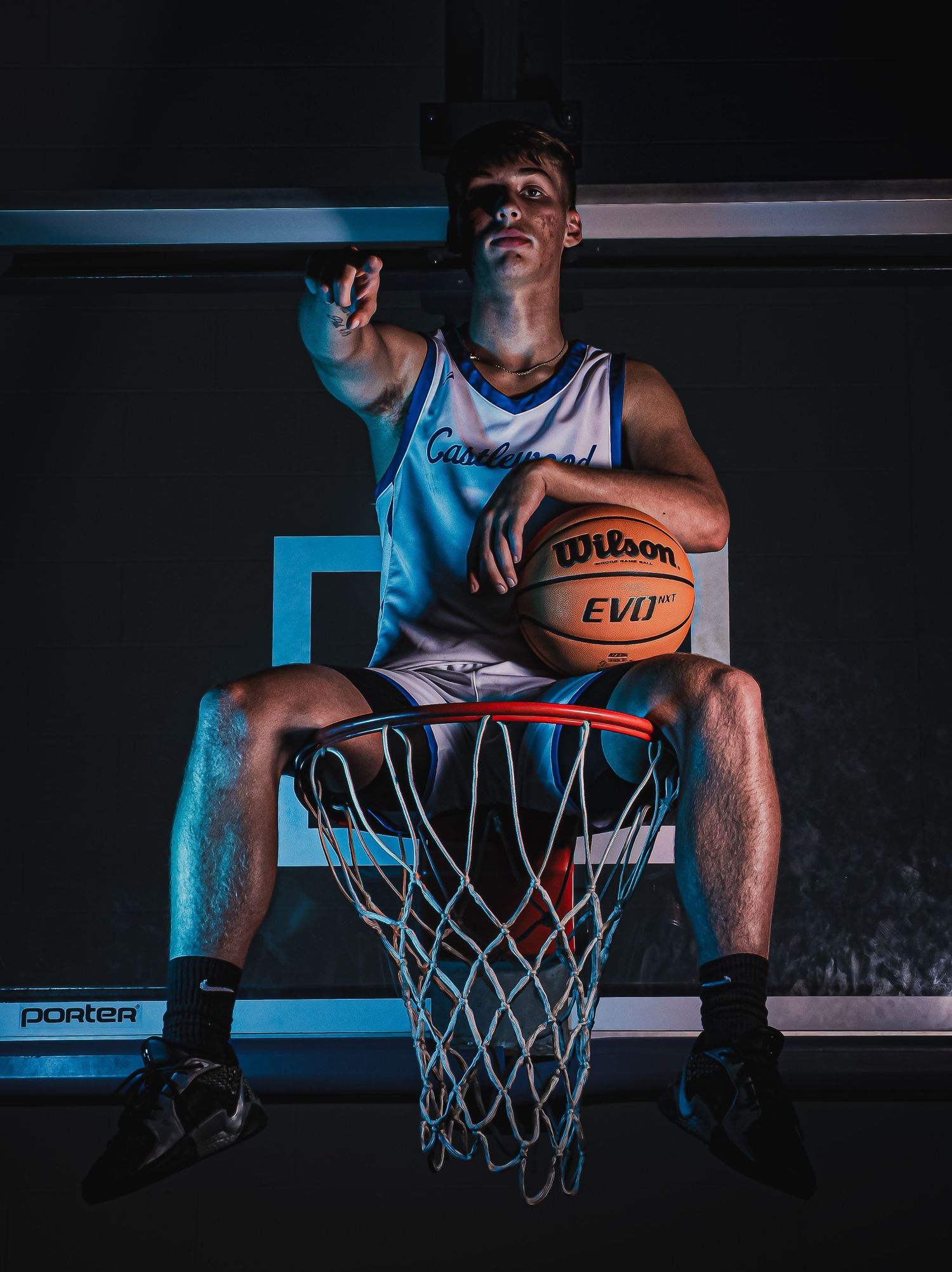 Basketball player sits on hoop holding Wilson basketball with blue lighting effect.