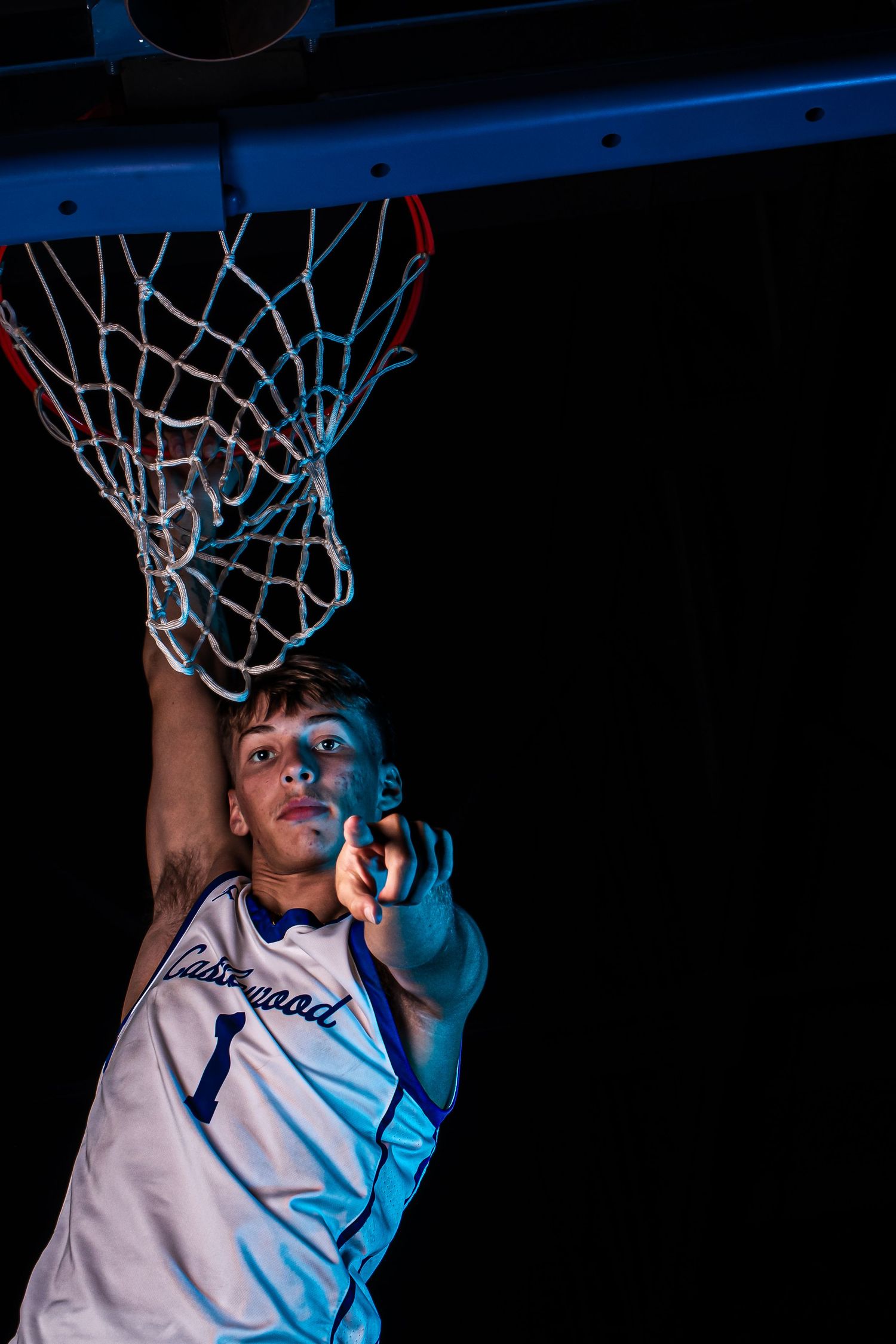 Basketball player in white jersey reaches up to touch net with dramatic blue lighting effect.