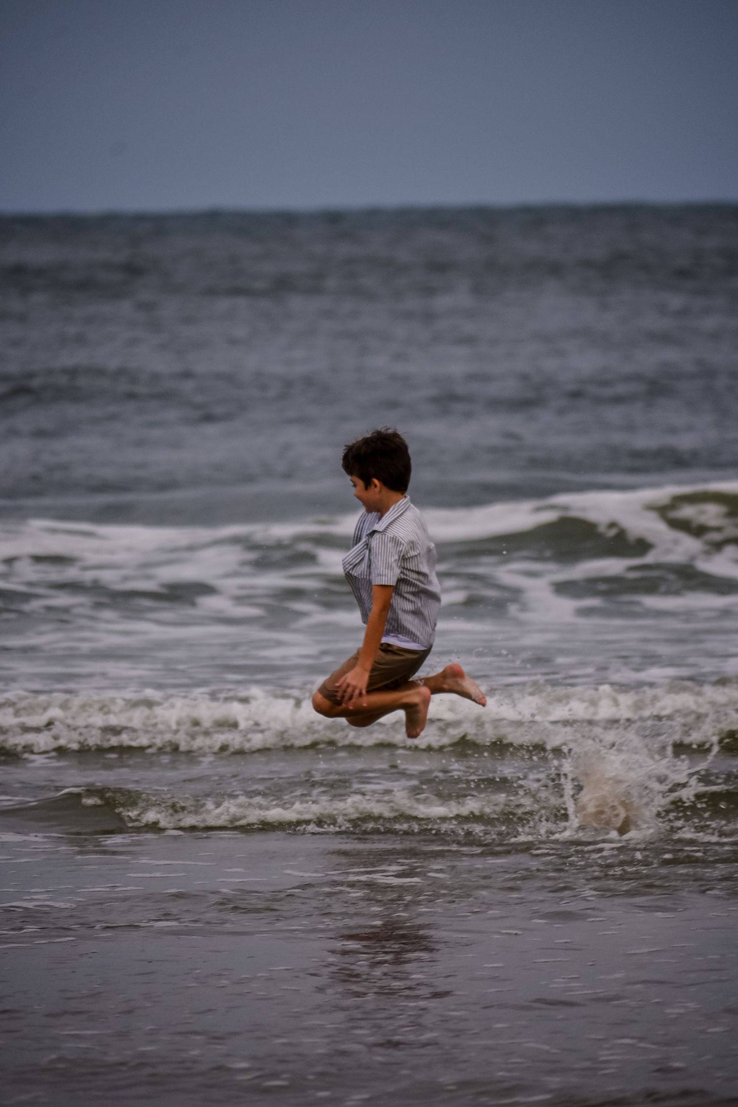 A young boy joyfully jumps over ocean waves on a beach, wearing a light shirt and shorts on an overcast day.