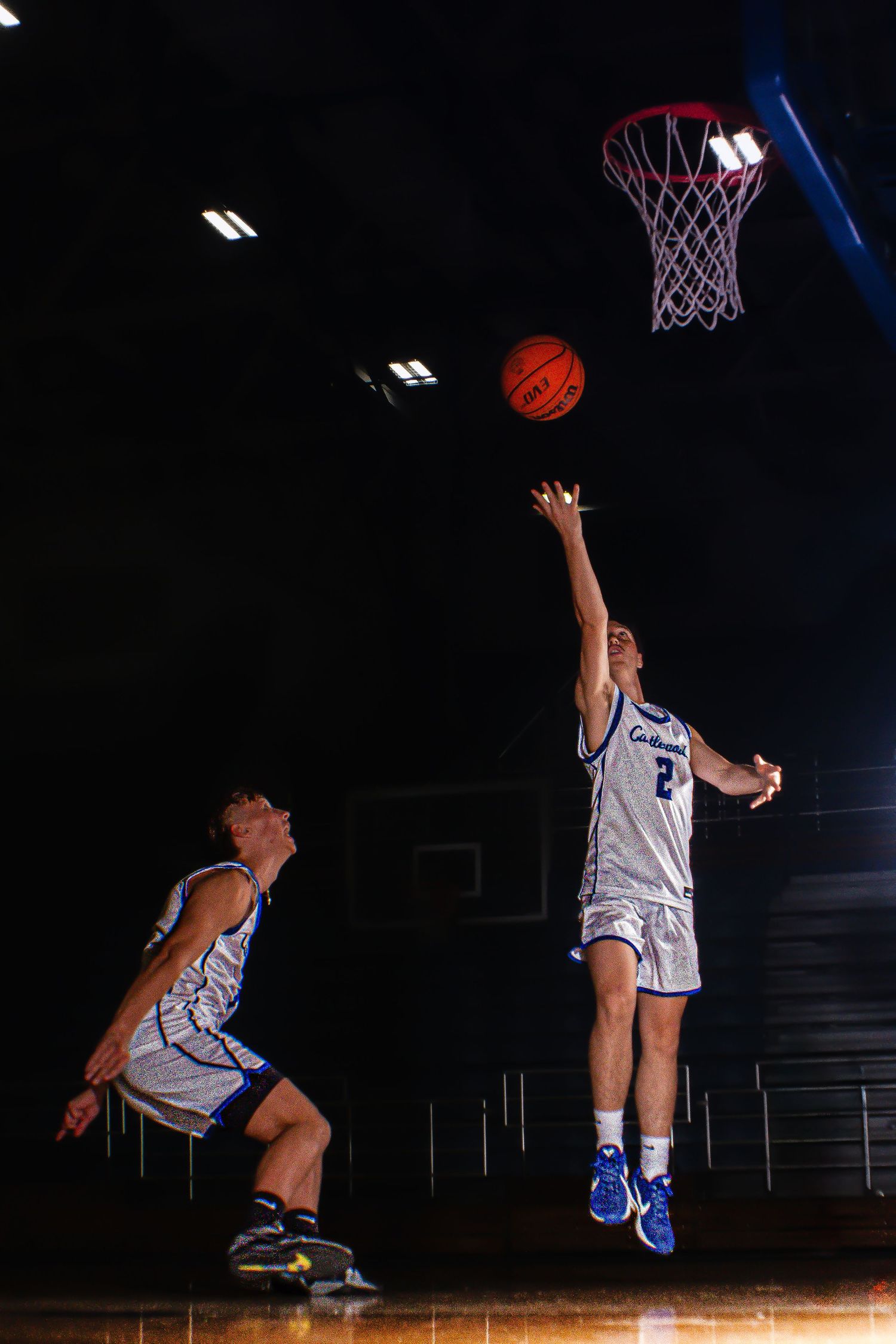 Two basketball players practice shooting drills in dimly lit gym.