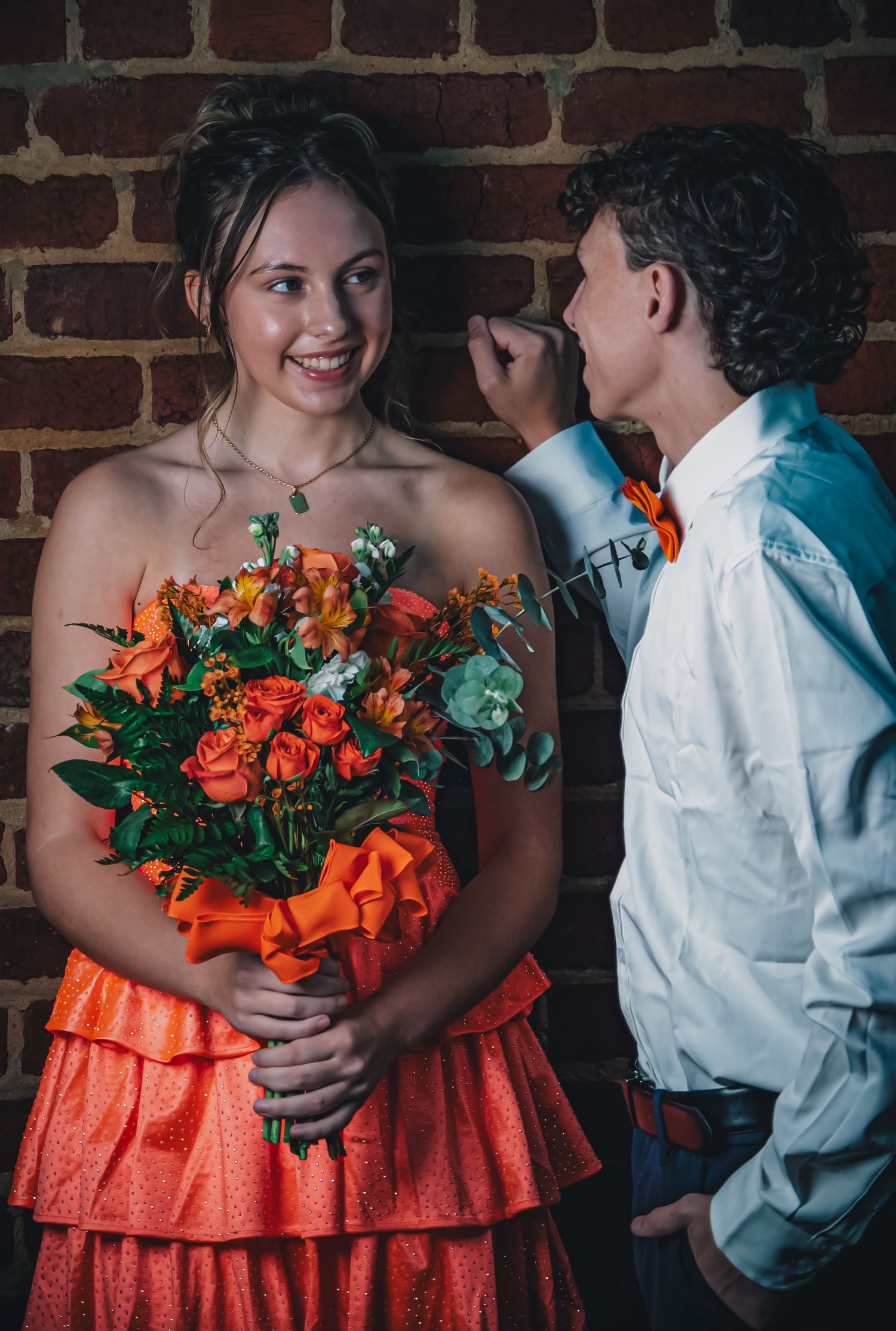 A romantic couple in coral and white embrace while holding a vibrant orange and green floral bouquet against a brick wall.