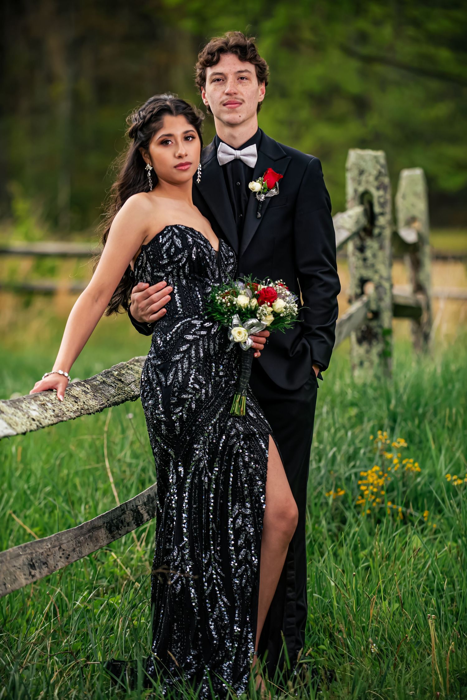A young couple in formal prom attire pose by a rustic fence, she in a black sequin gown, he in a black tuxedo with red boutonniere.