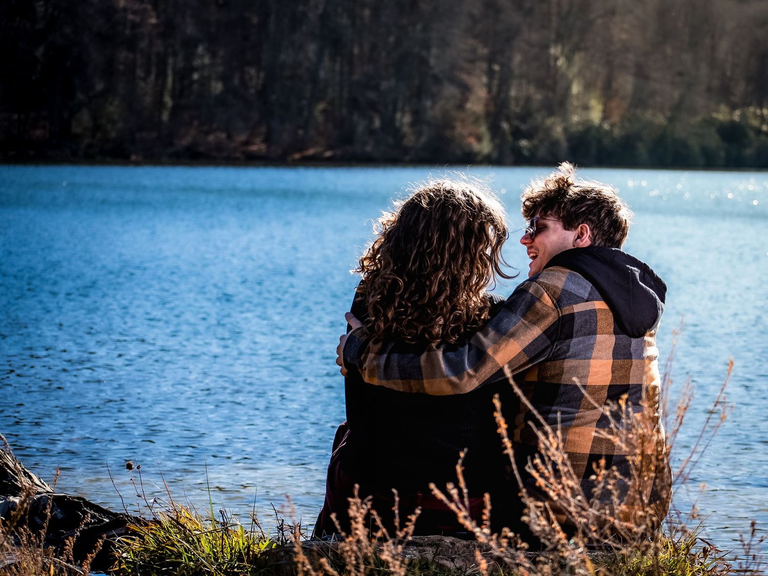 Two people sitting close together by a serene lake surrounded by autumn grass and trees at sunset.