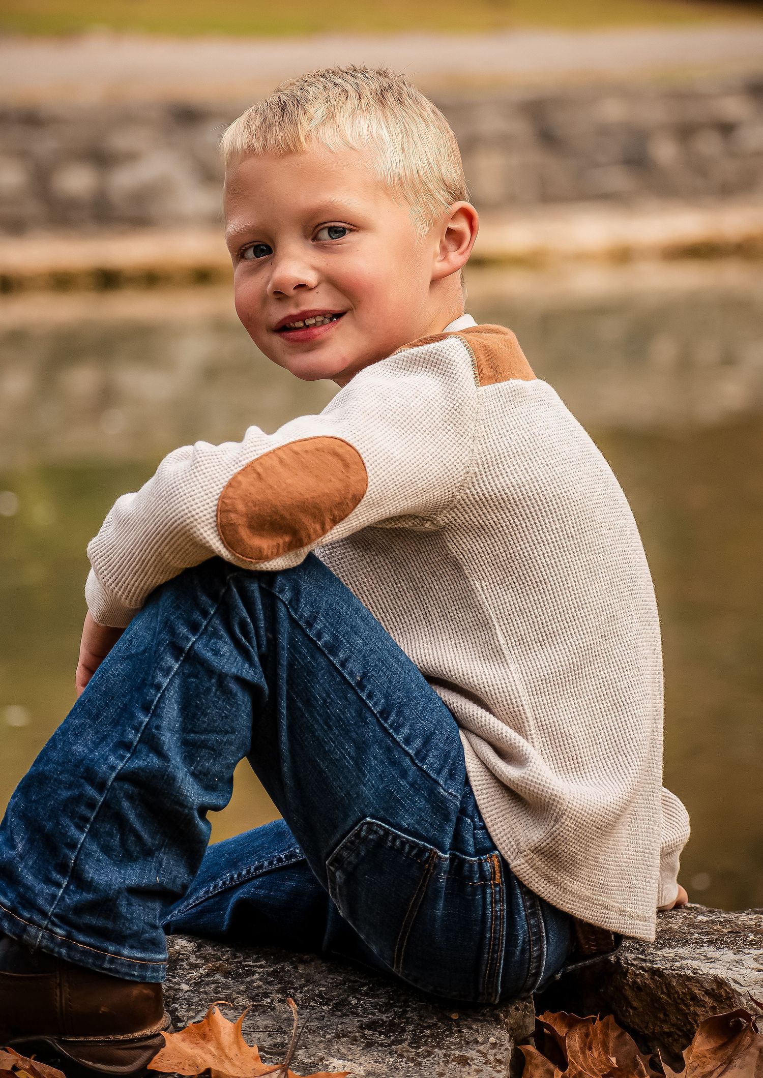 A young child in blue jeans and a beige sweater sits casually on a stone wall near water.