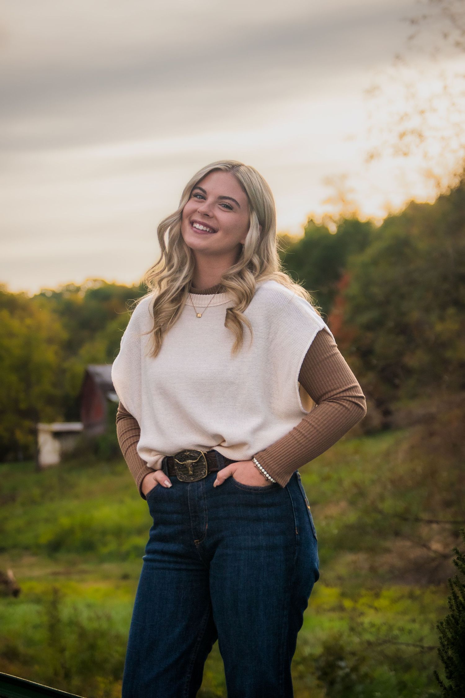 Person in white sweater and jeans smiling outdoors during golden hour.