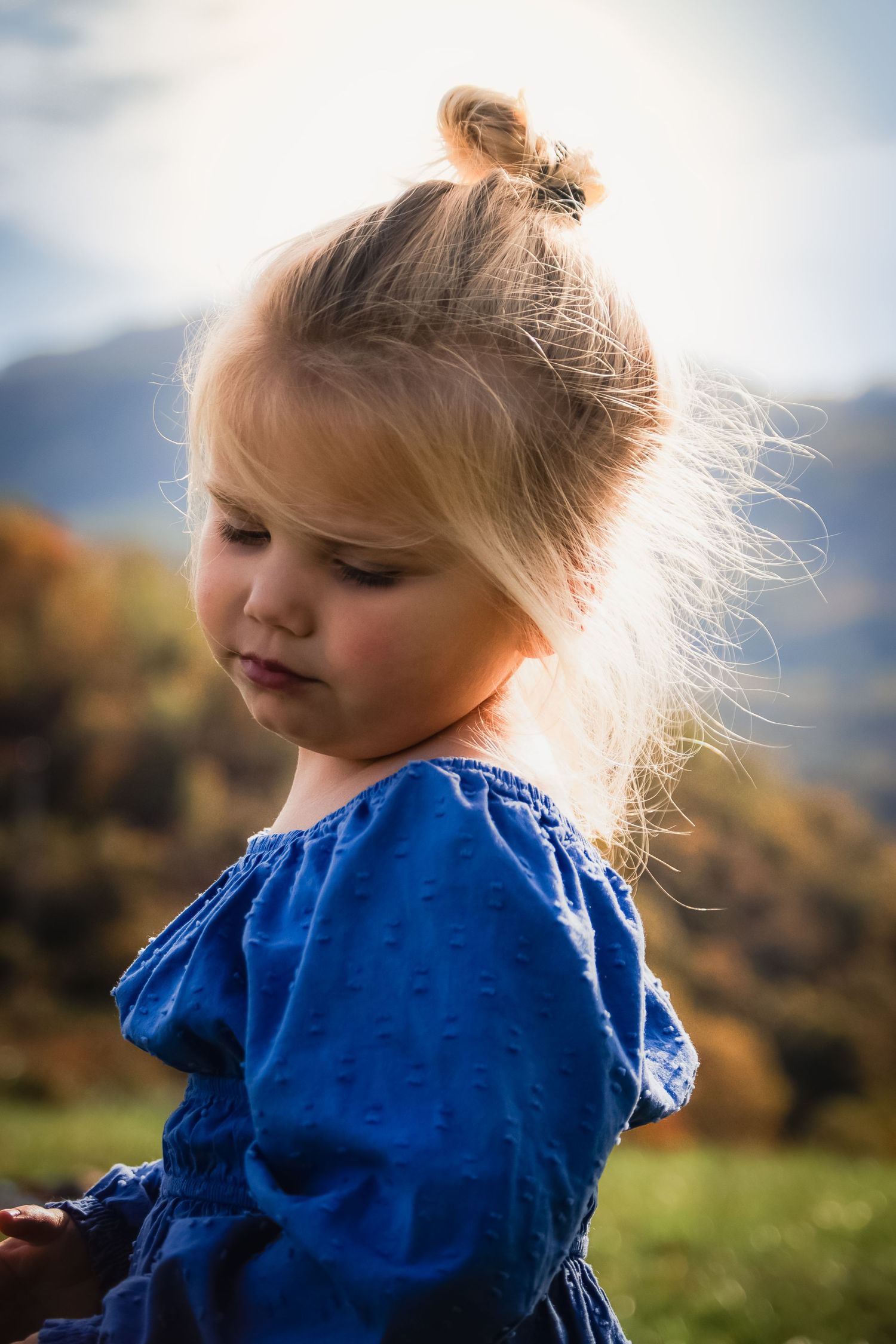 Child in a blue dress with hair in a bun standing outdoors against mountains and autumn foliage.