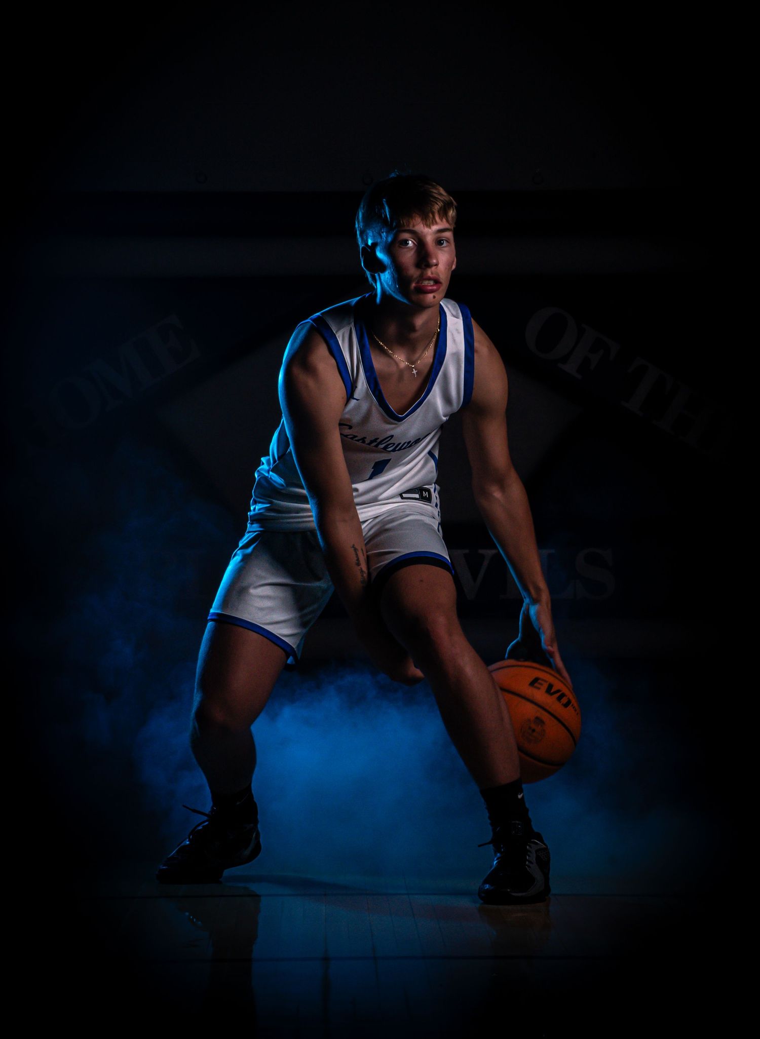 Basketball player in white uniform crouches low on dark court with dramatic blue lighting.