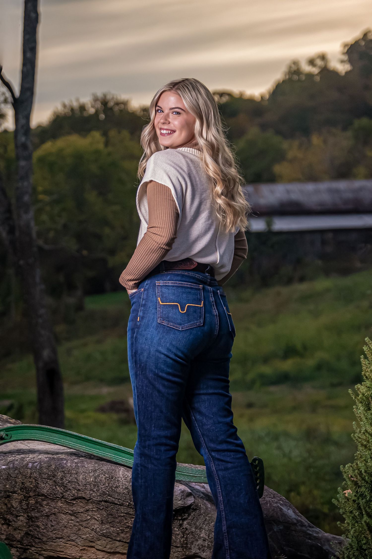 Person in casual outfit poses on rustic fence during sunset in countryside setting.