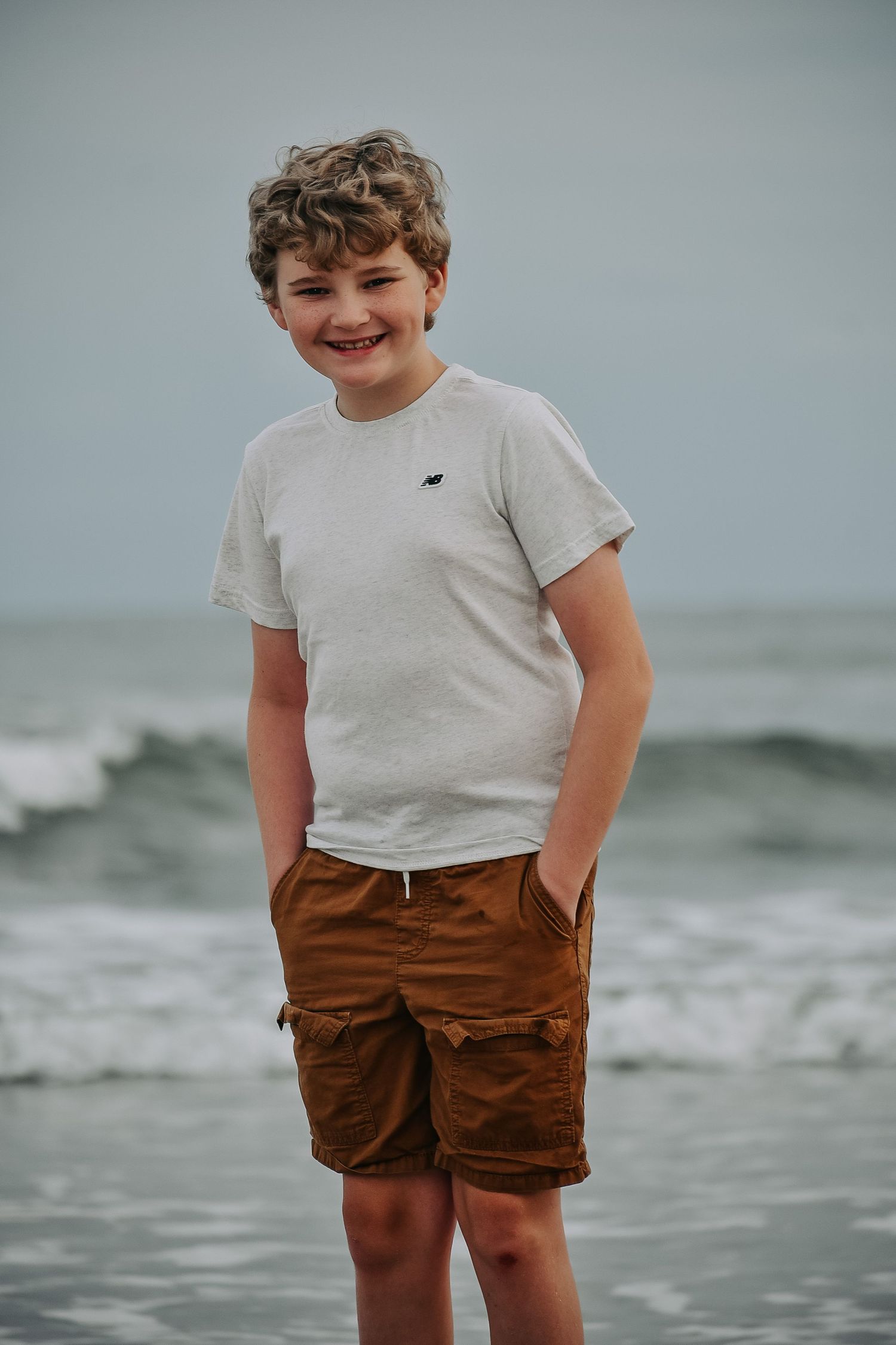 Young person in casual attire smiling while posing on beach with waves in background.