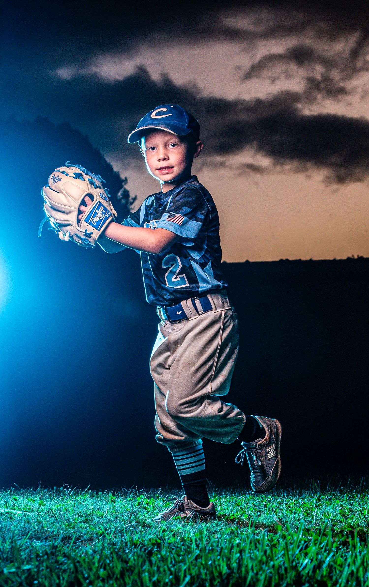 Young baseball player in blue uniform poses dramatically at sunset against moody sky with dramatic lighting effects.