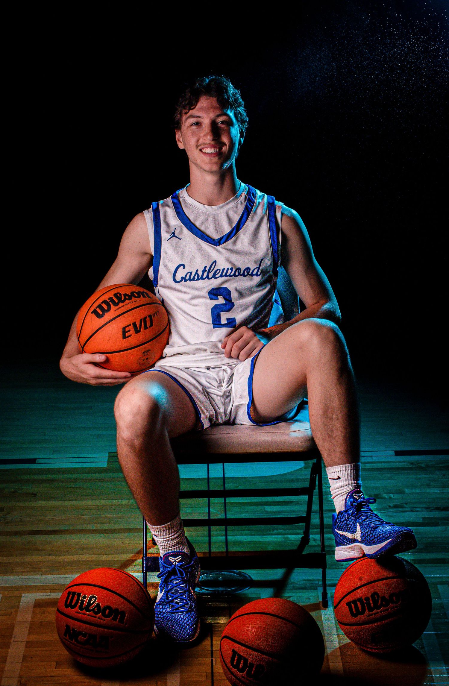 Basketball player poses with multiple Wilson basketballs in Castlewood jersey on wooden court.