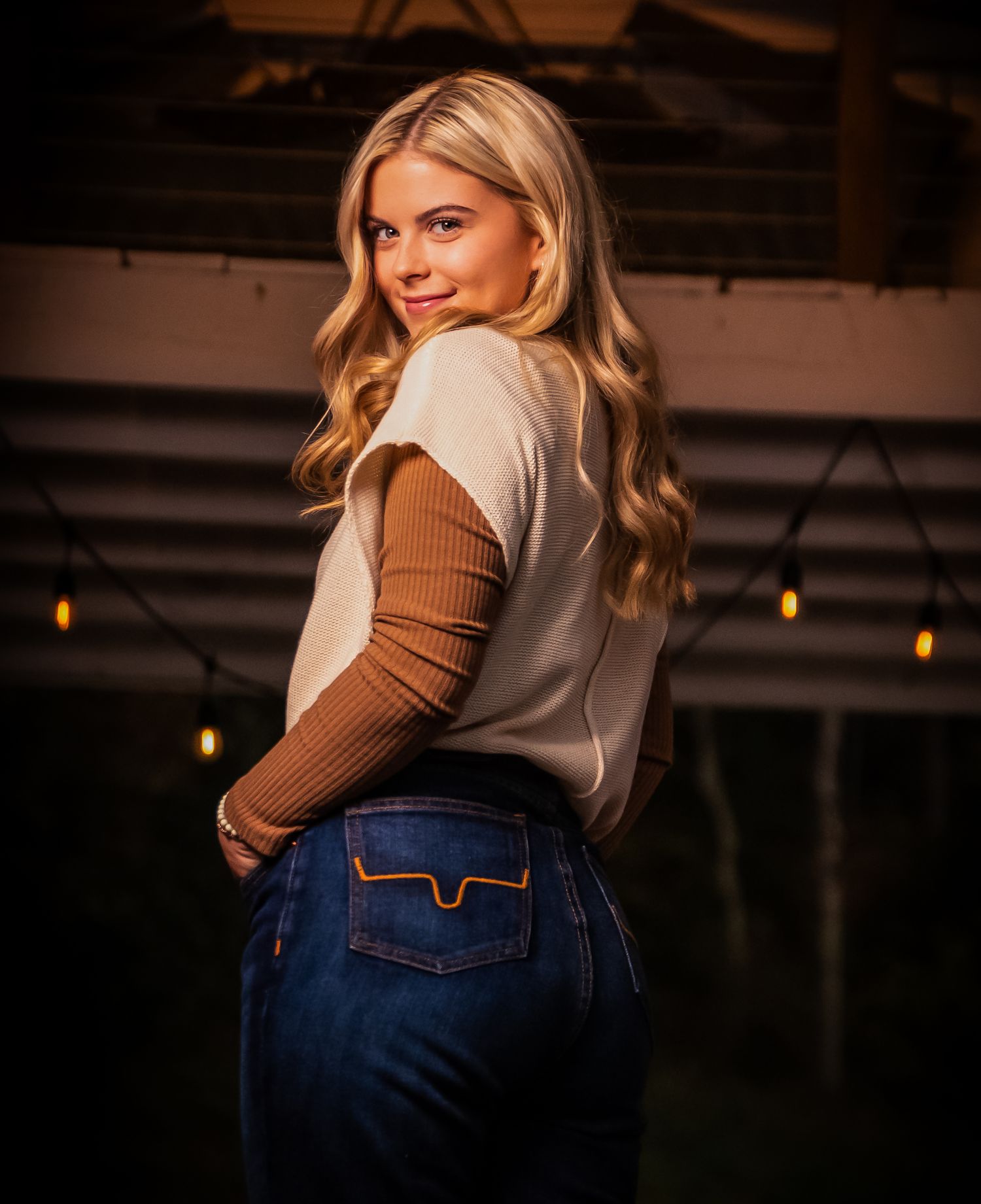 Woman in beige sweater vest and brown long sleeve top poses against dark background.