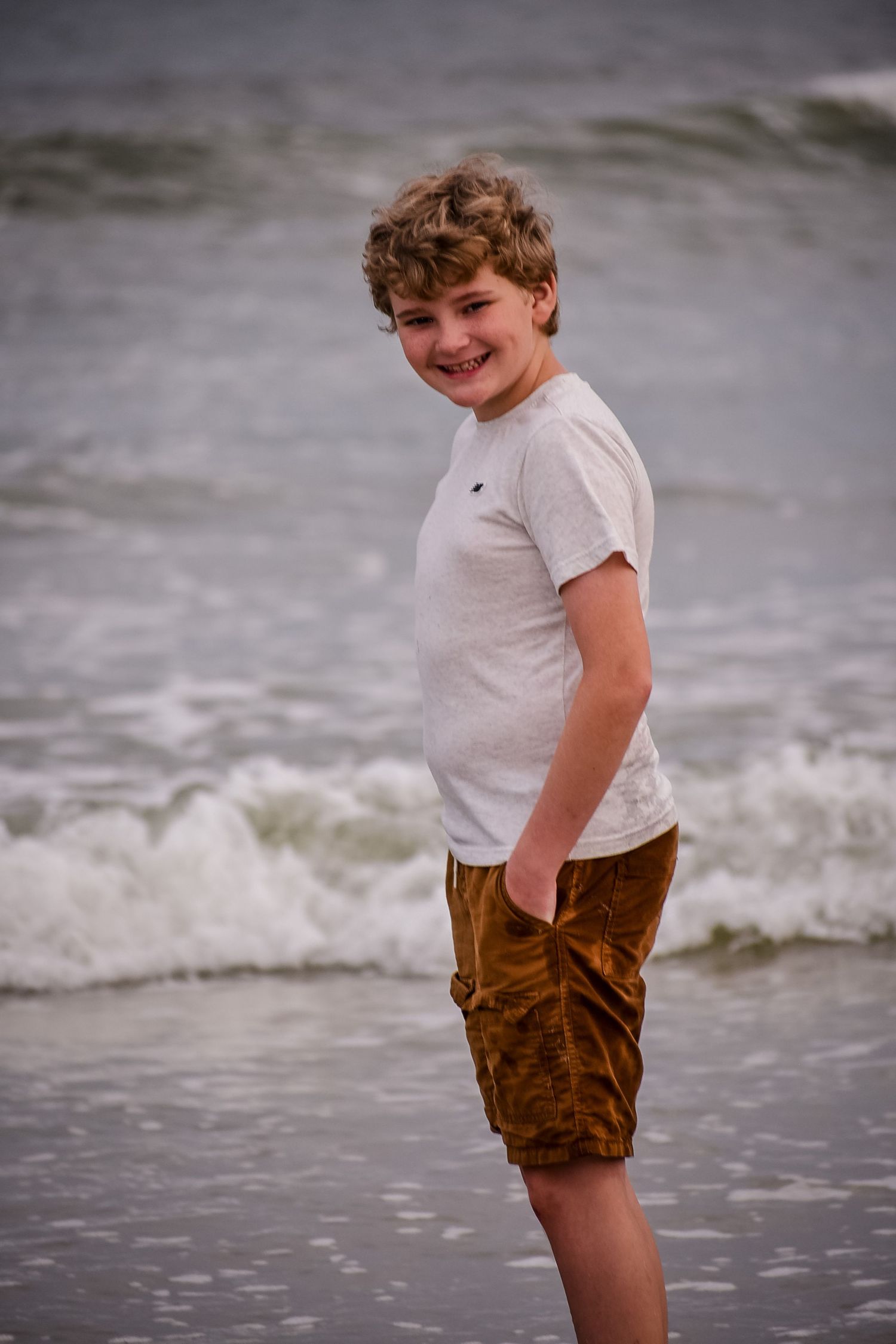 Smiling boy in white t-shirt and brown shorts standing on a sandy beach with ocean waves in the background.