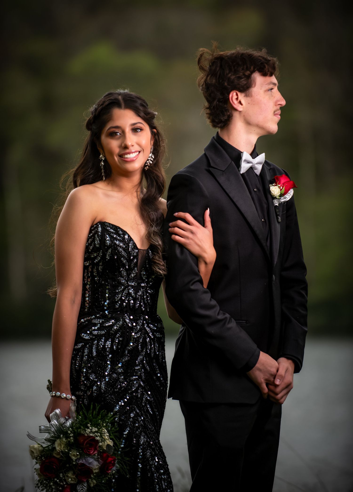 A young couple dressed in formal prom attire, she in a black sequin gown, he in a black tuxedo with red boutonniere.
