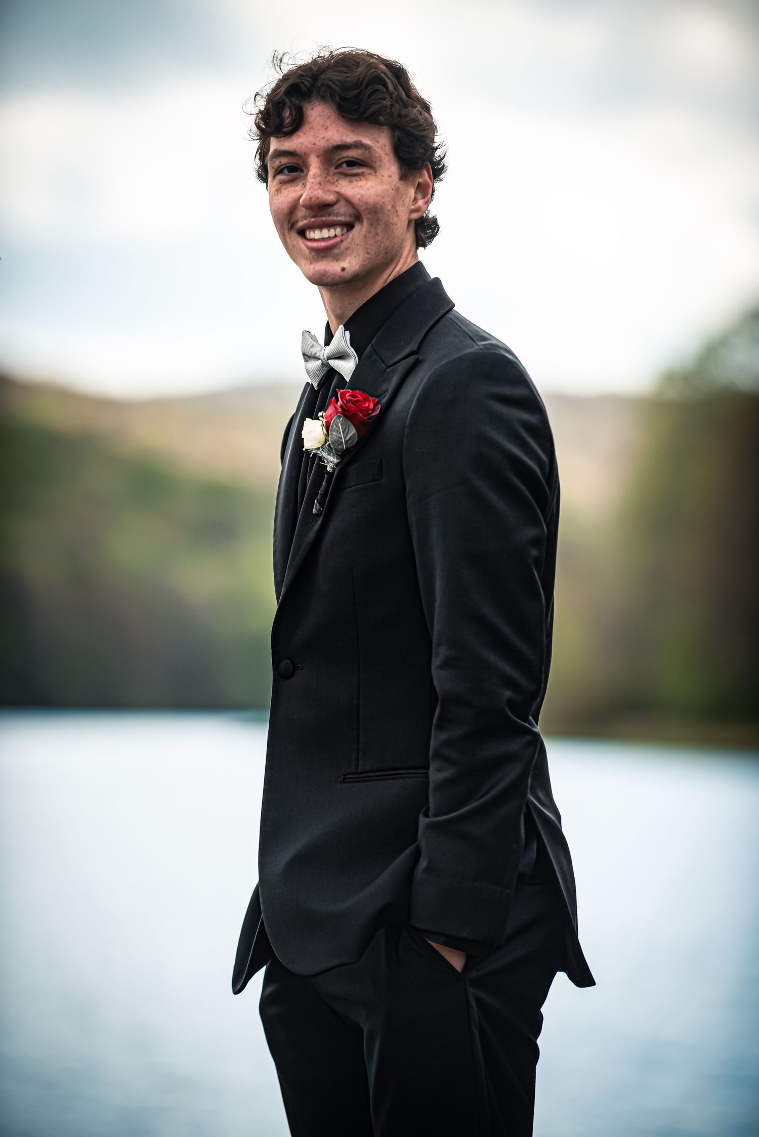 Smiling young man in black tuxedo with bow tie and boutonniere posing outdoors near a lake.