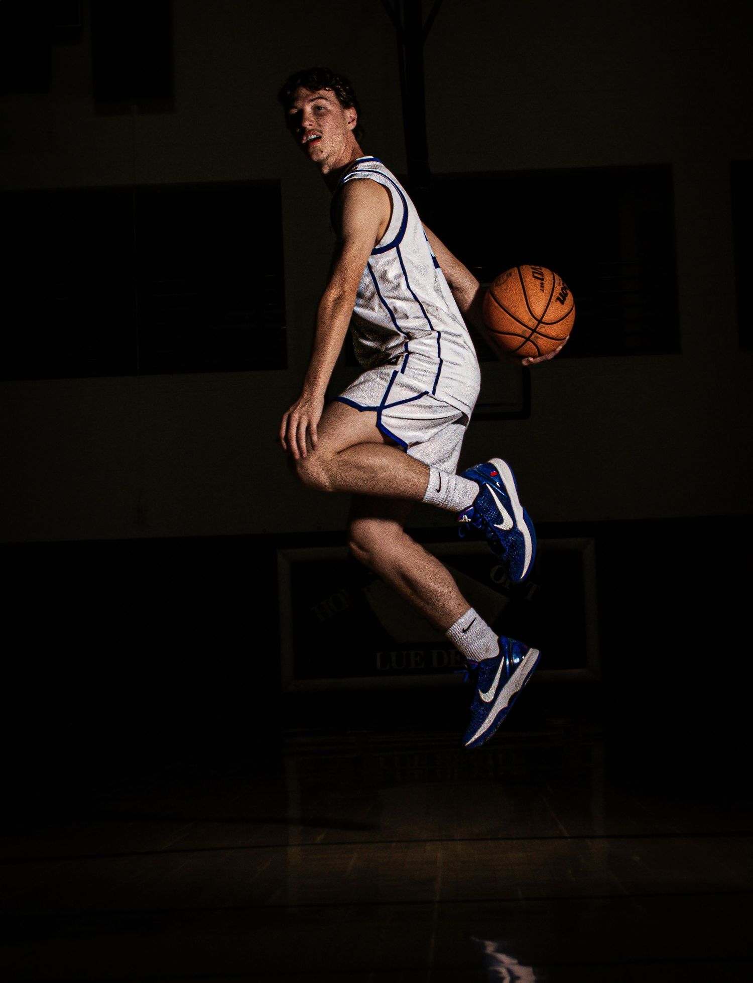 Basketball player performs mid-air move while holding ball in dark gymnasium.