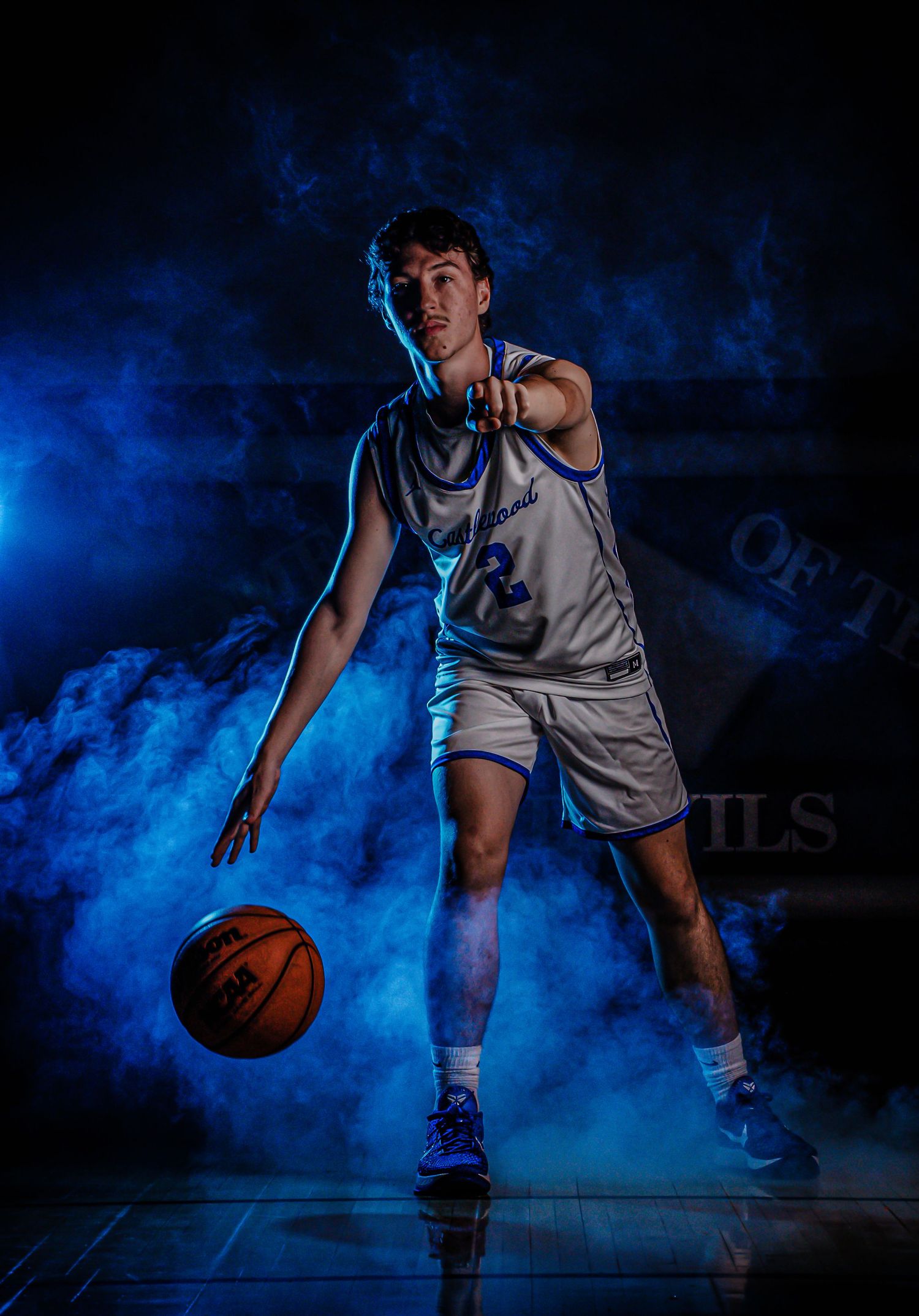 Basketball player performs dribbling moves through blue smoke effects on dark court.