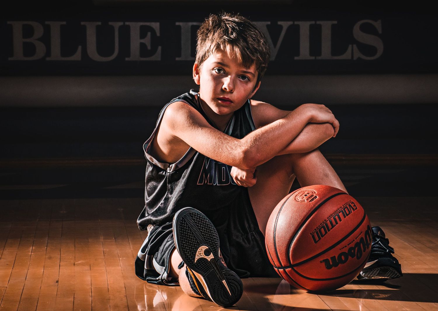Youth athlete in black uniform sits on wooden court with basketball in moody lighting.