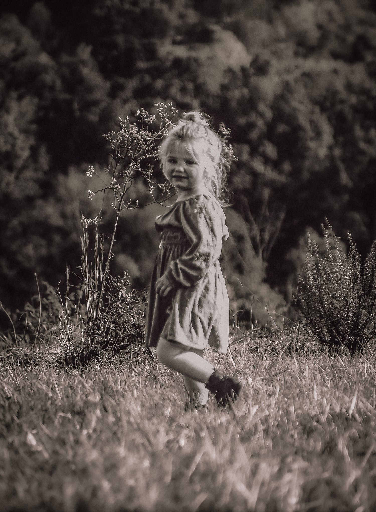 A black and white artistic portrait of a person sitting in a field of tall grass and wildflowers.