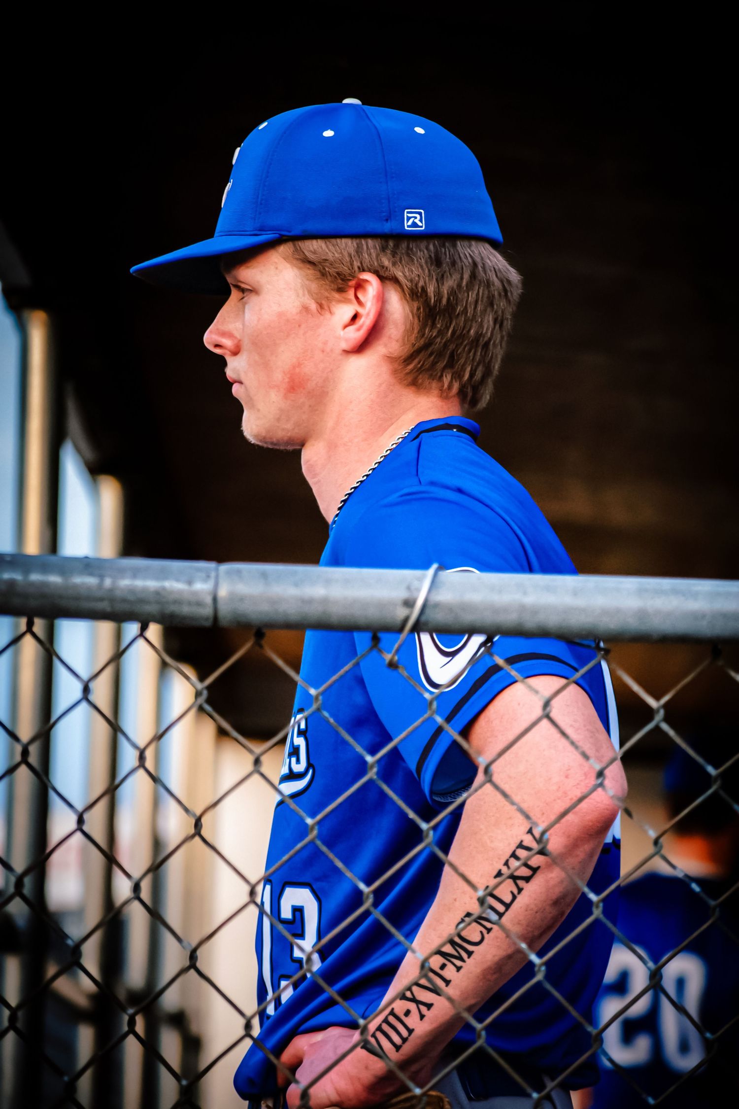 Baseball player in blue uniform and cap stands by chain-link fence dugout, showing Roman numeral tattoo on forearm.