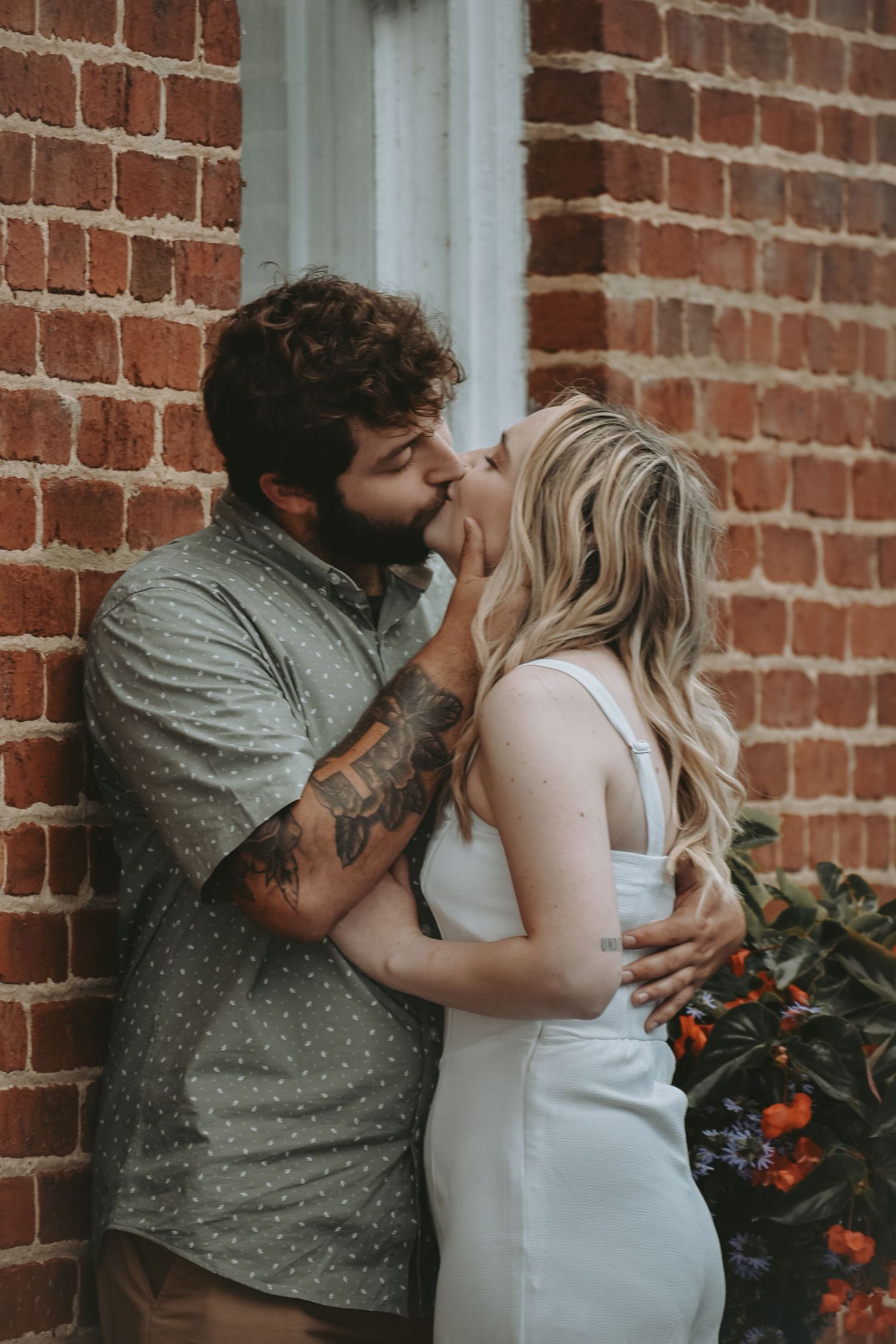 Romantic couple poses against rustic brick wall with flowering plants.