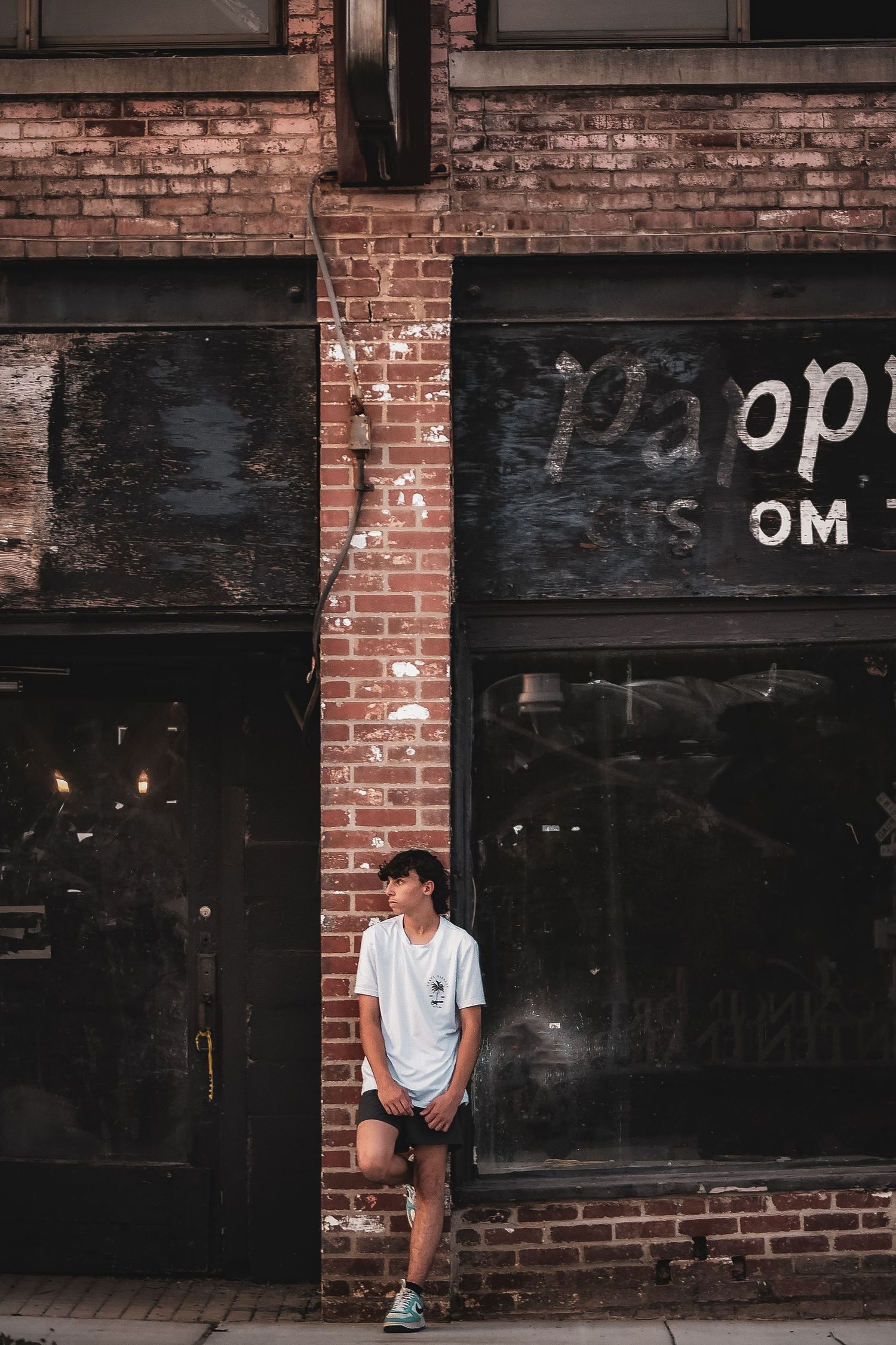Person in white shirt sits against storefront with black panels and brick walls.