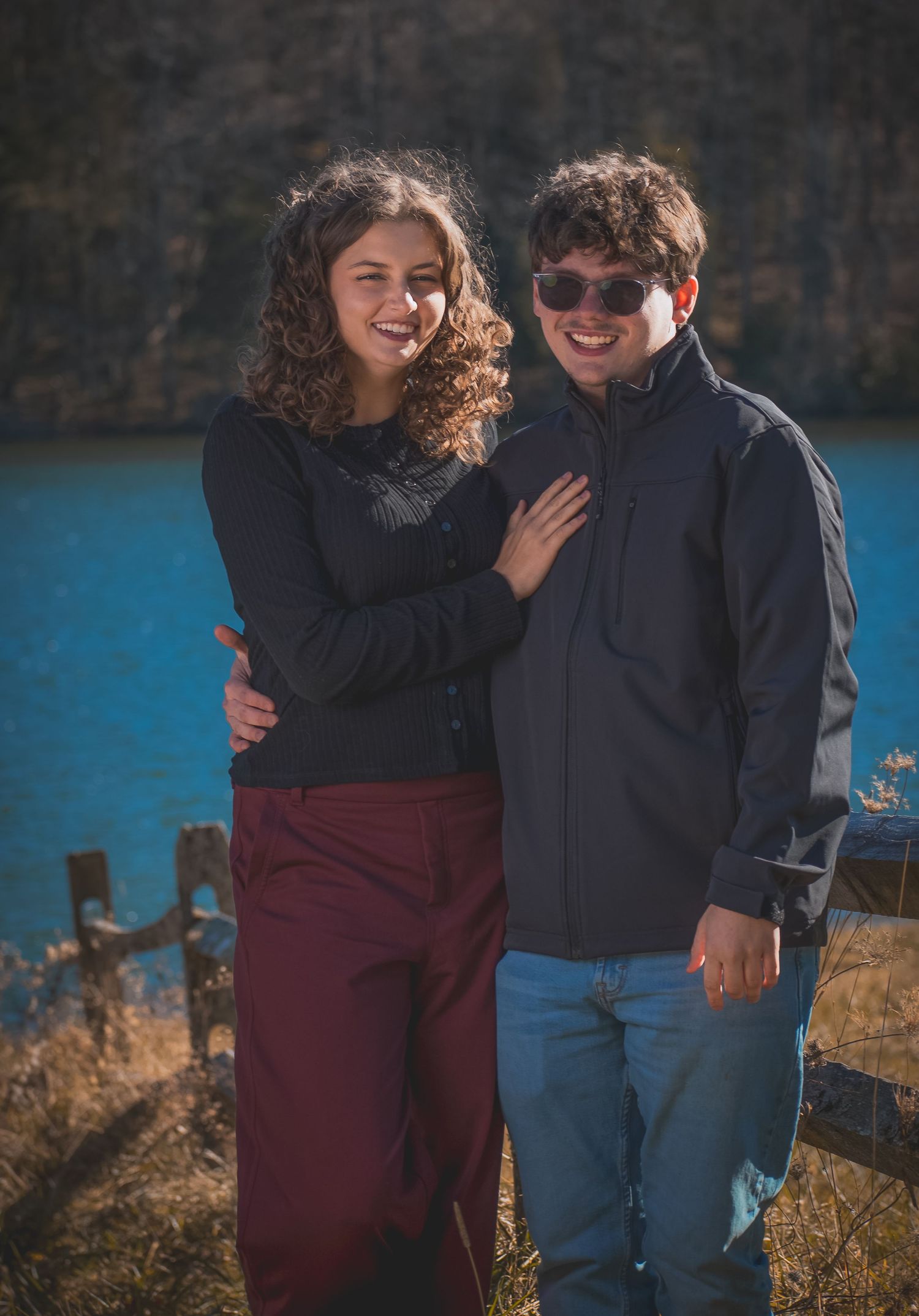 A couple shares joyful laughter while standing together against a scenic blue lake backdrop on a sunny day.