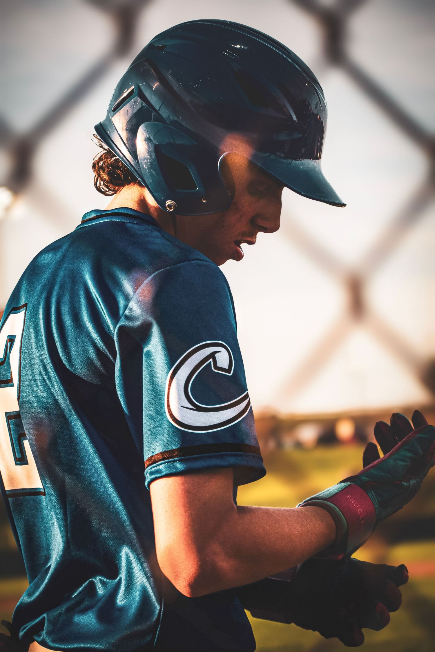 Young baseball player in teal jersey and batting helmet adjusting gloves near chain-link fence.