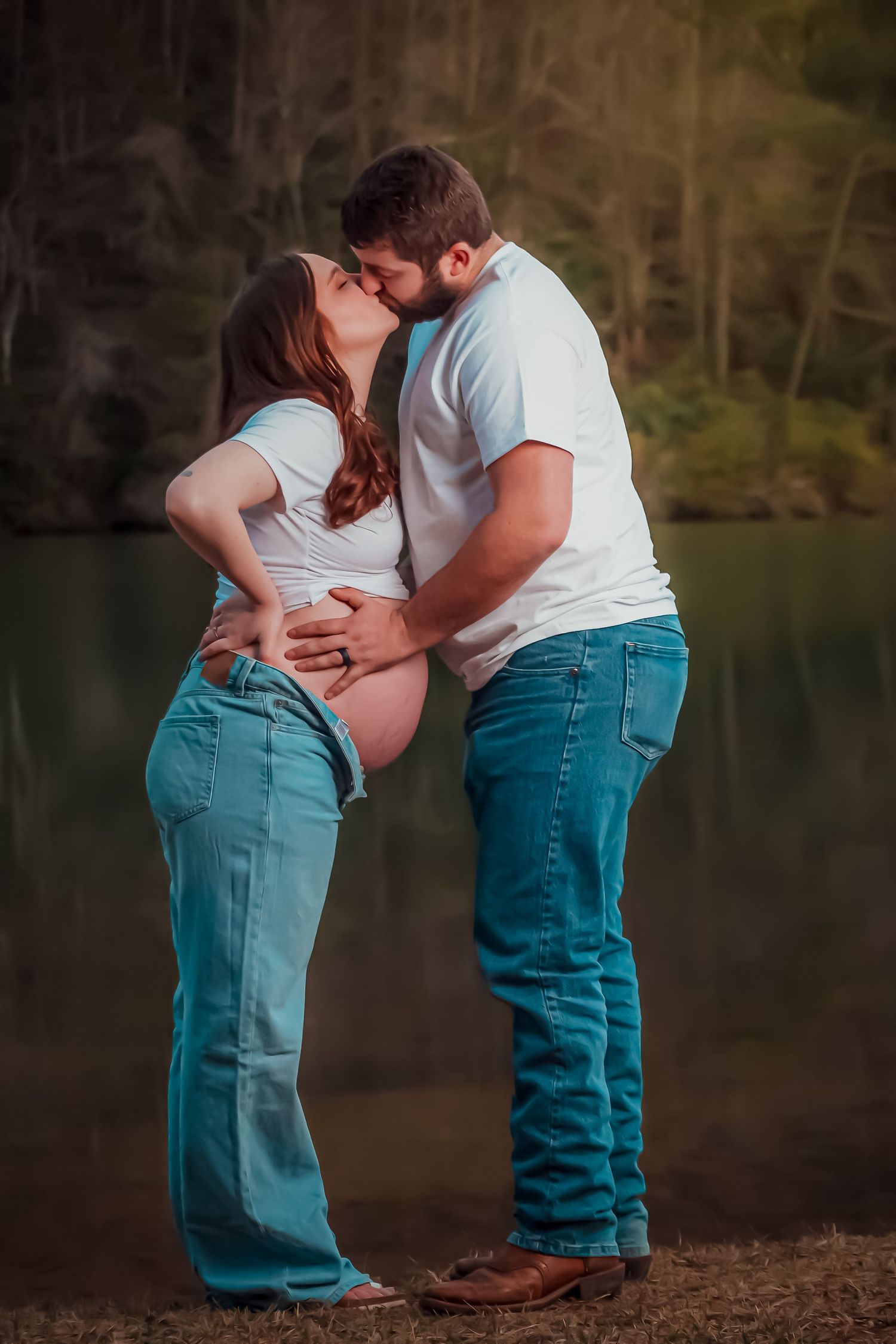 A couple in matching jeans and white shirts share a romantic moment by a serene lake at sunset.