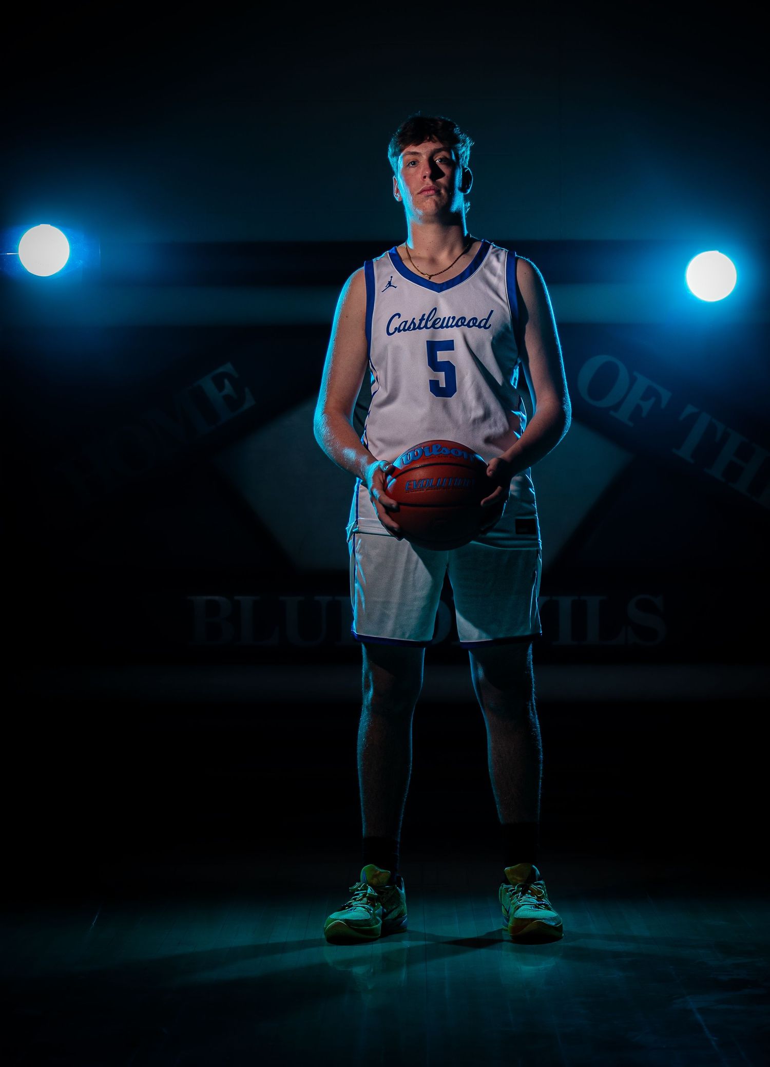 Basketball player stands in dramatic lighting holding ball on dark court with glowing blue spotlights in background.