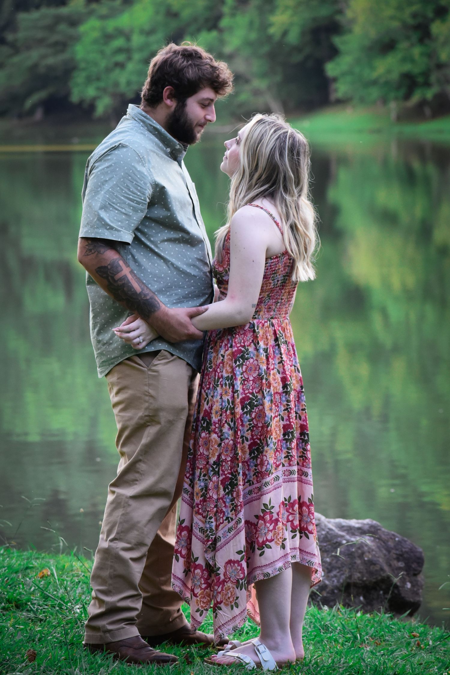 A tender moment between a couple standing near a peaceful lake surrounded by greenery.