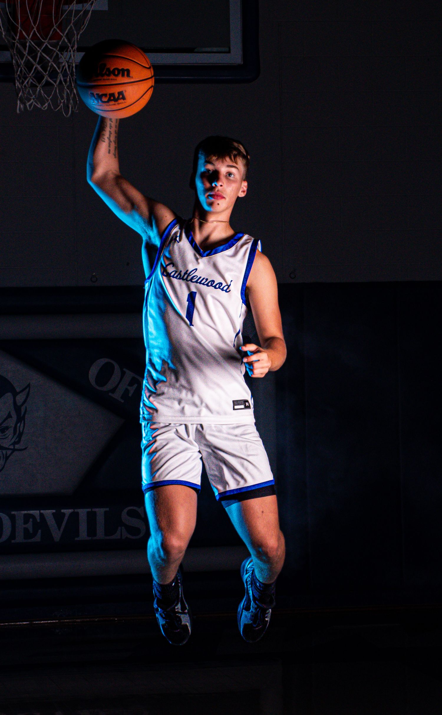Basketball player in white uniform jumps with ball against dark background with blue lighting.