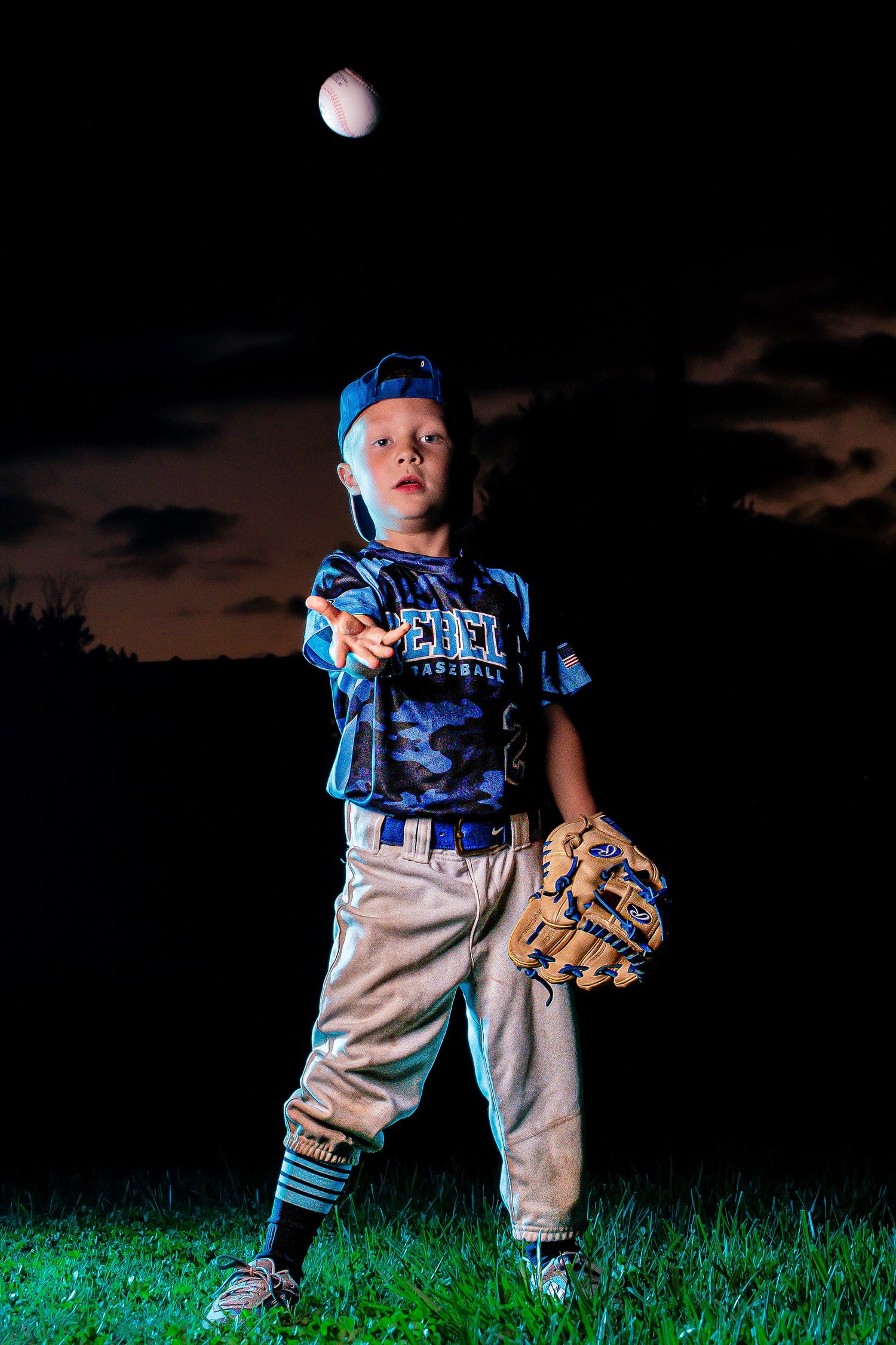 Young baseball player in blue uniform poses with glove against dark sky with moon.