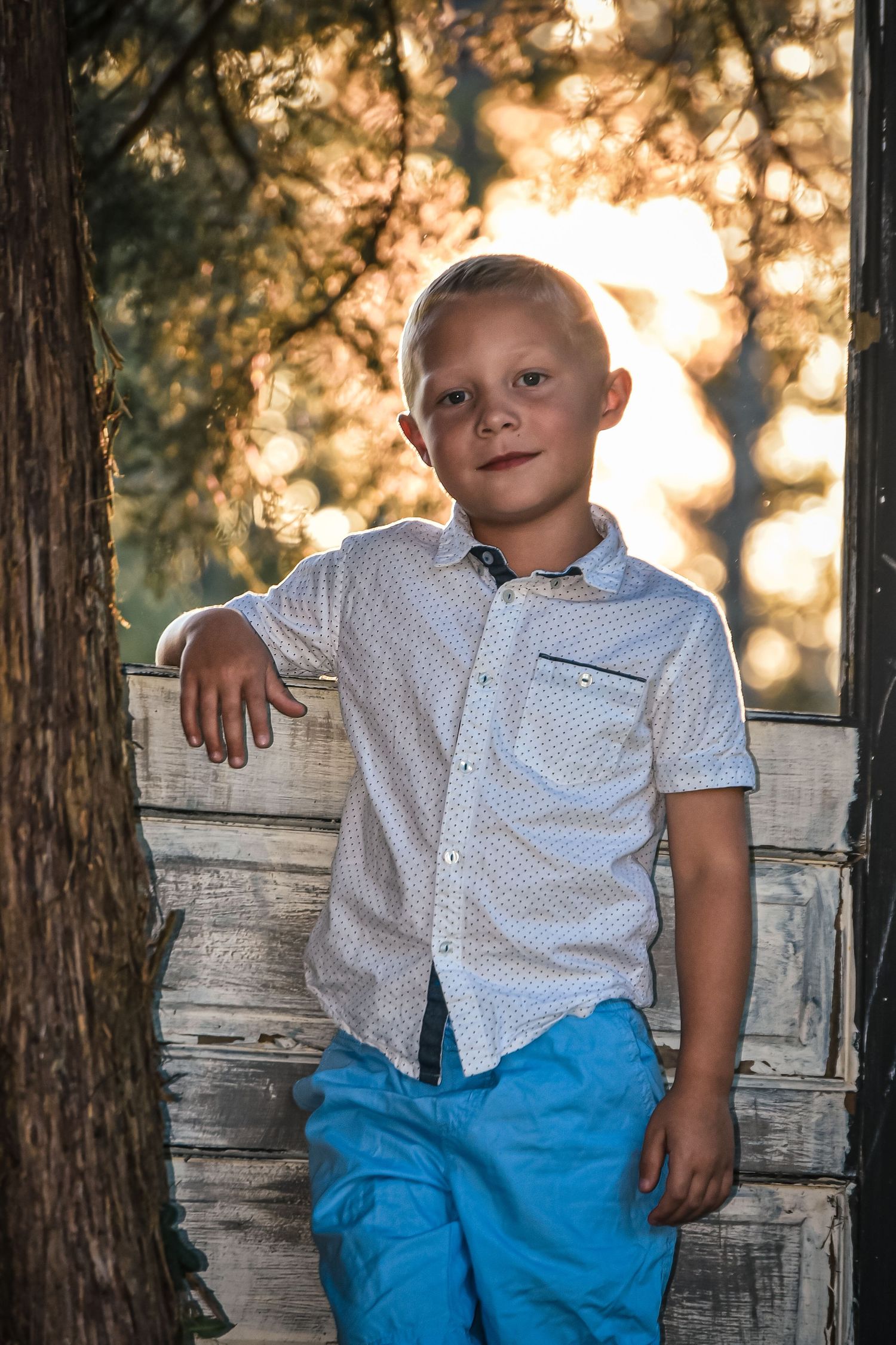Young child in polka dot shirt and blue shorts sitting on rustic wooden bench.