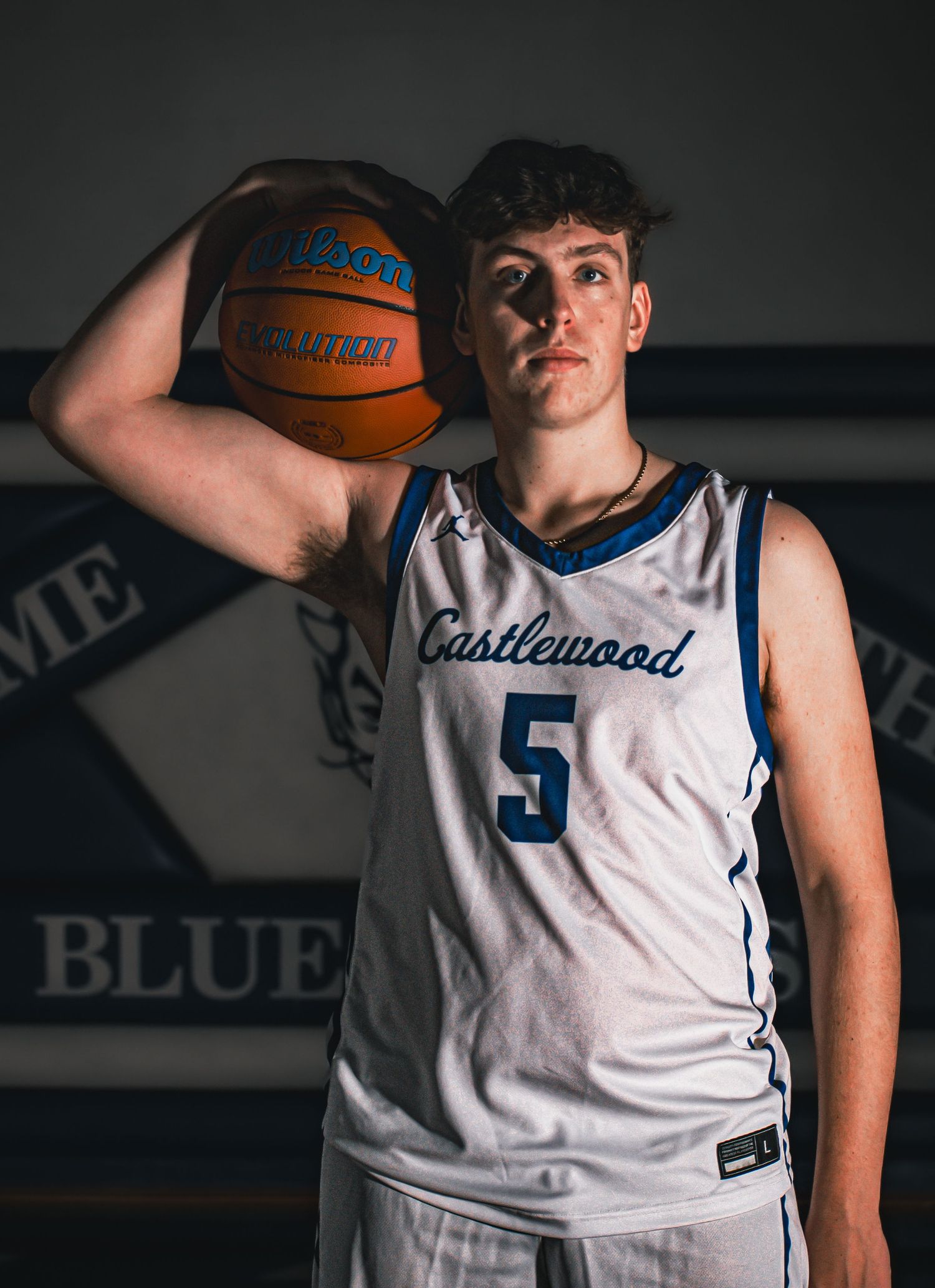 Basketball player in white Castlewood jersey number 5 poses with ball in dramatic lighting against dark background.