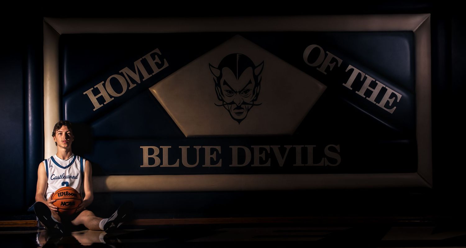 Basketball player sits beneath large 'Home of the Blue Devils' sign in dramatic lighting.