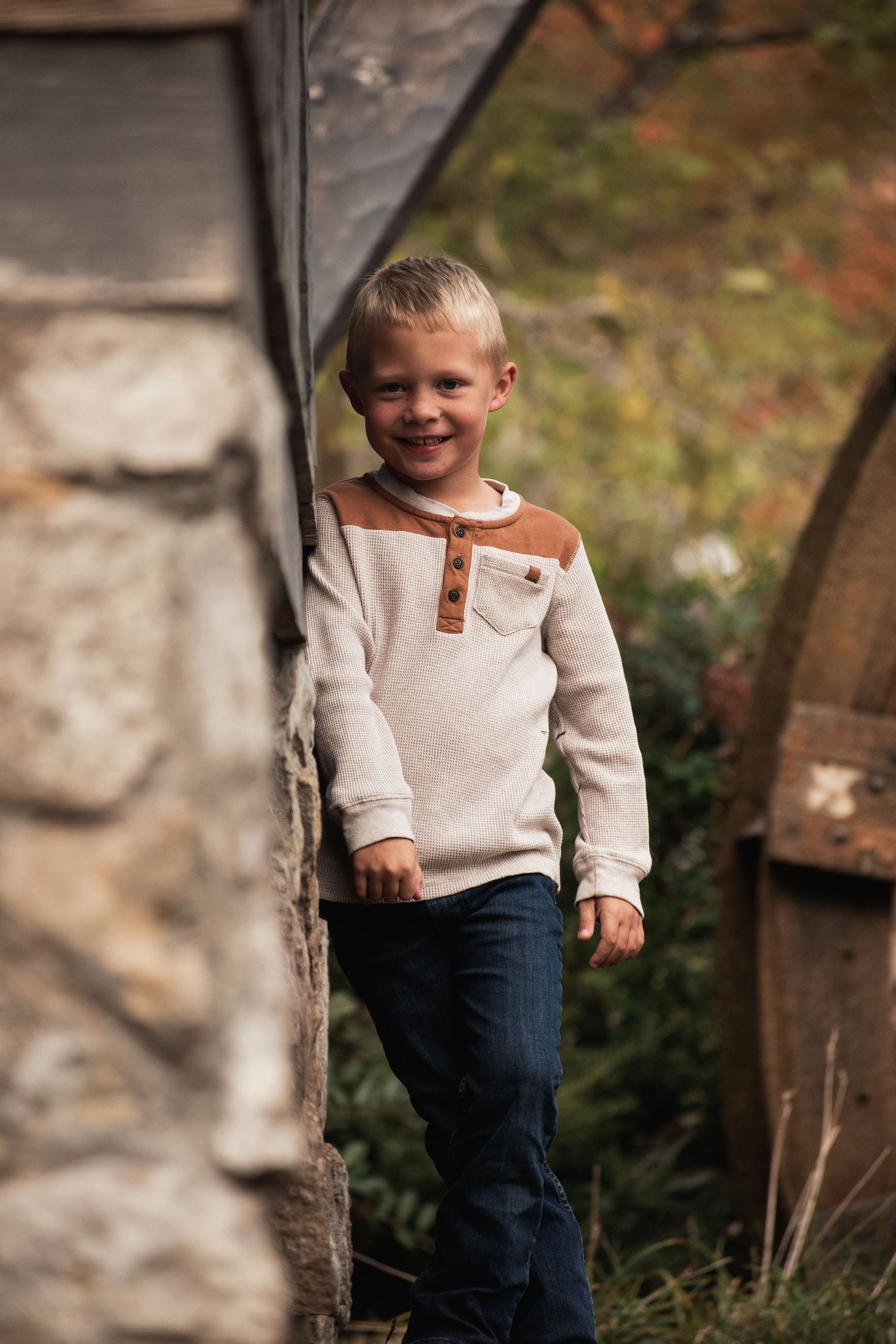 A young child in a cream henley shirt smiles while standing against a stone wall.