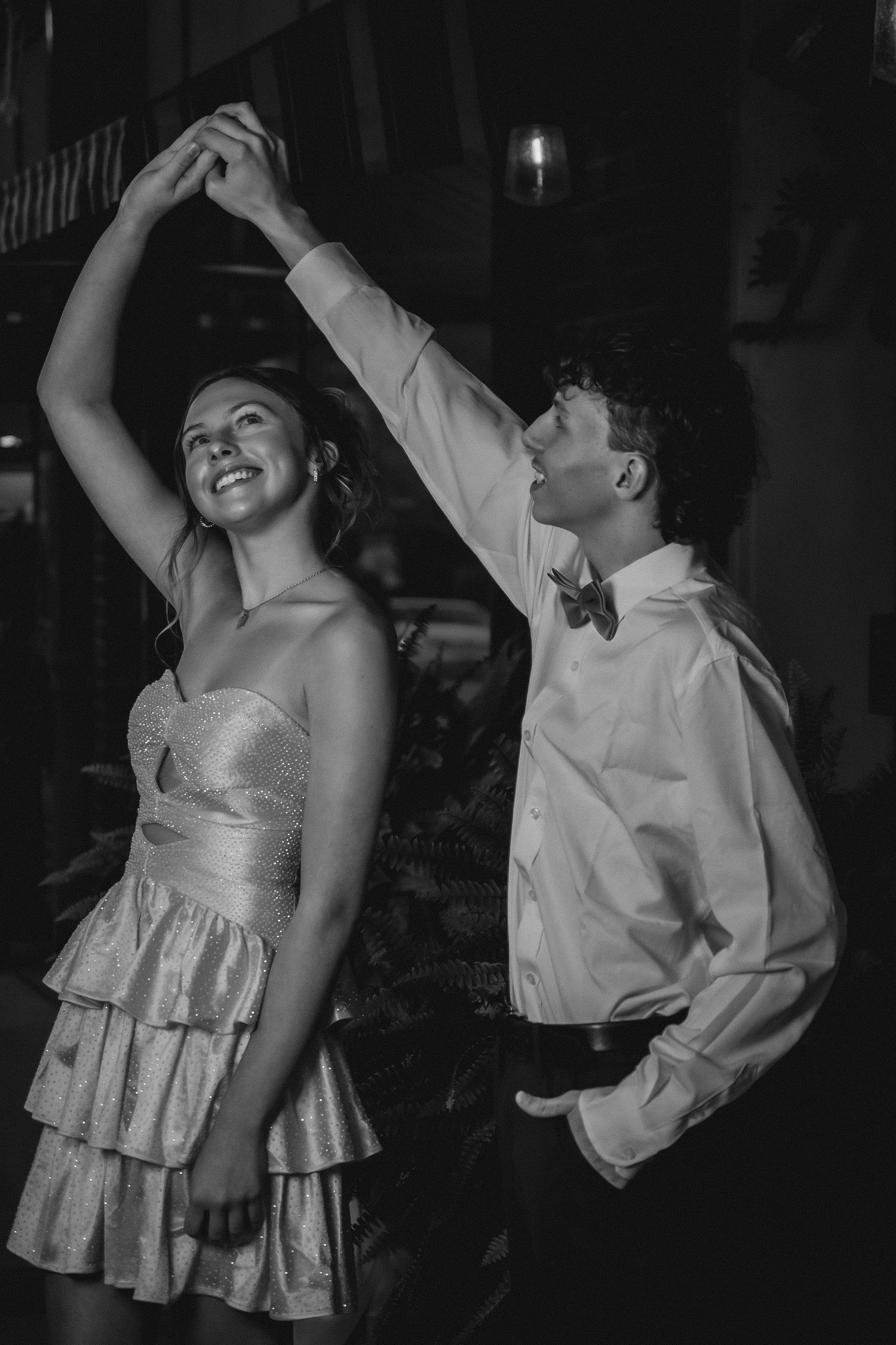 A couple dances together at a formal event, with the woman in a tiered dress and man in a white shirt and bowtie.