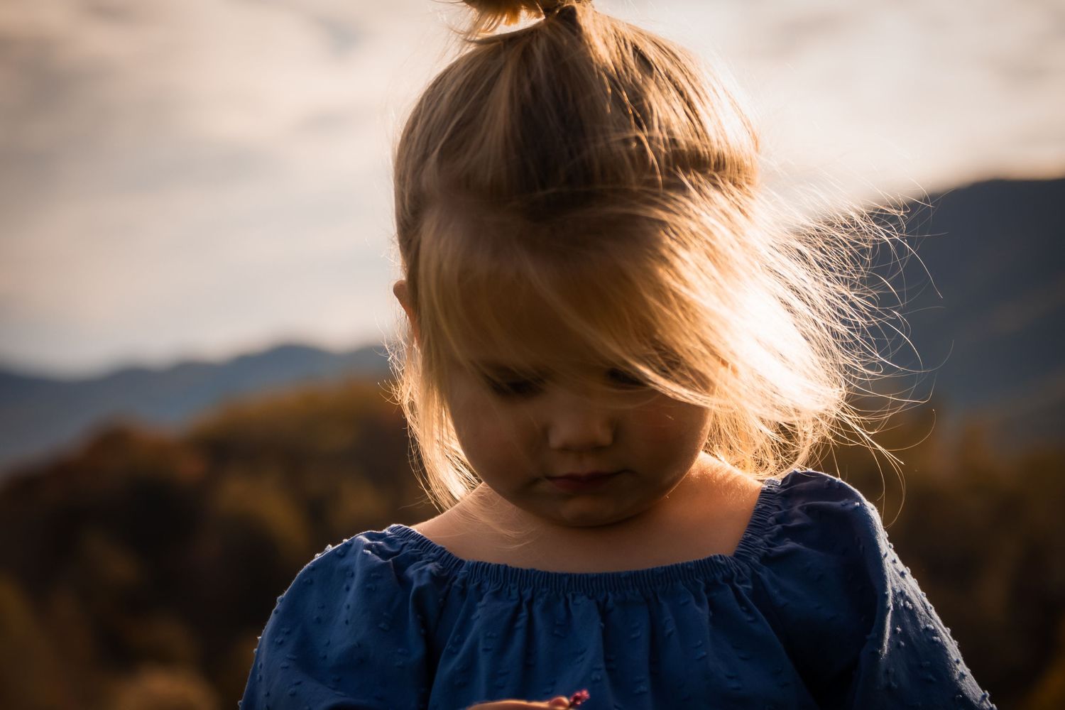 Young child in blue dress backlit by golden sunset in mountain landscape.