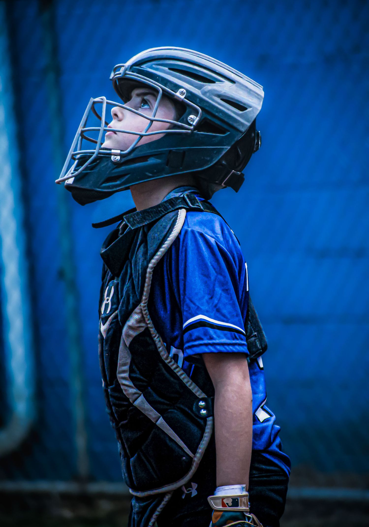 Young baseball catcher in blue jersey and protective gear with helmet and chest guard looks upward.