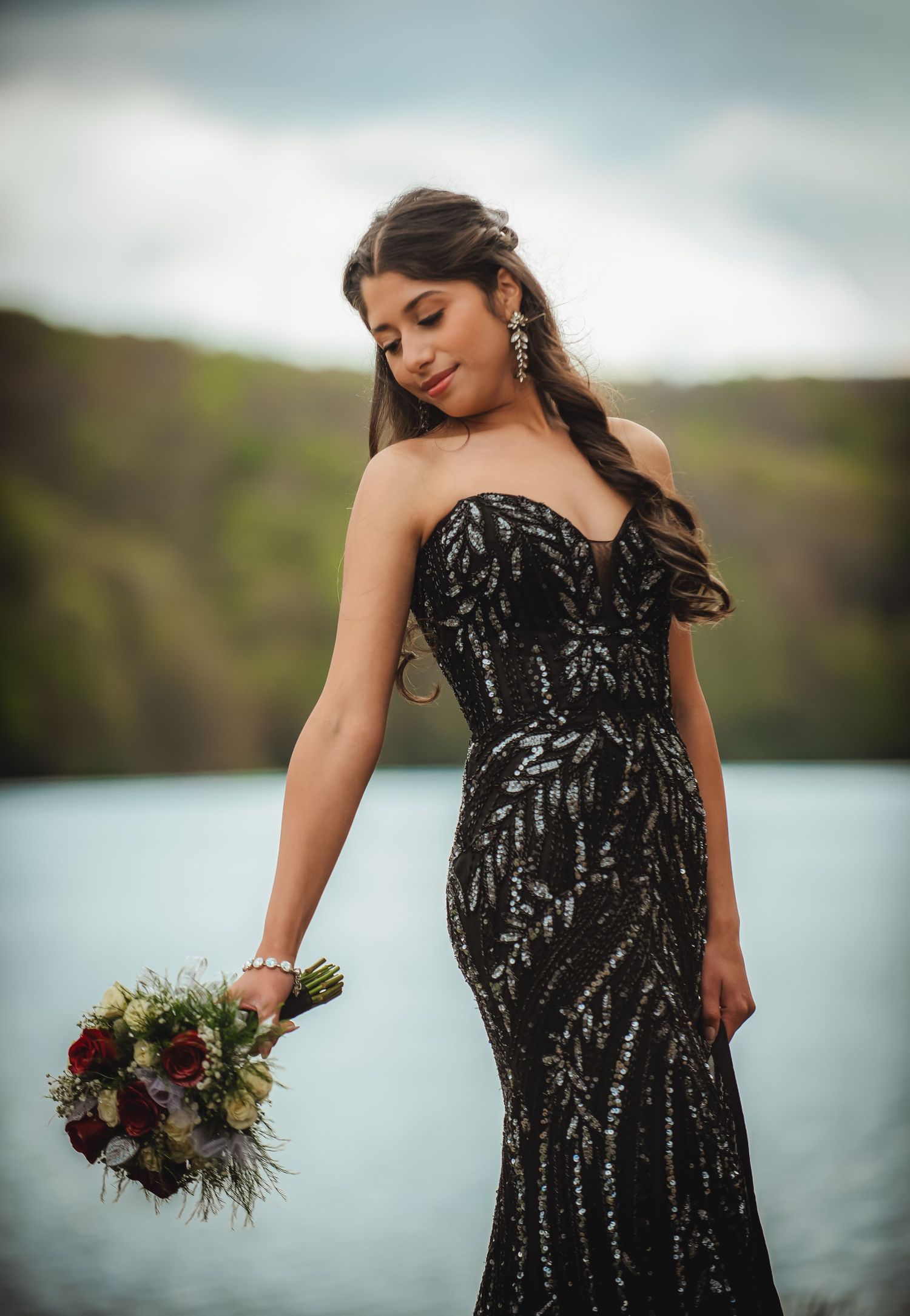 Woman in black sequin strapless gown holding bouquet by lakeside.
