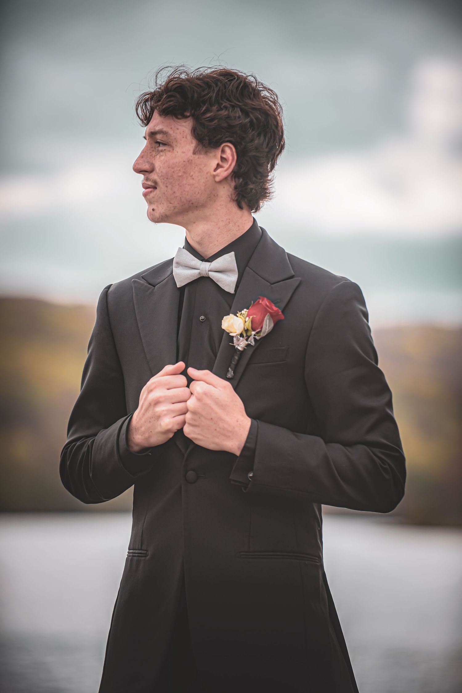 Young man in black tuxedo with gray bow tie and boutonniere poses outdoors at formal event.