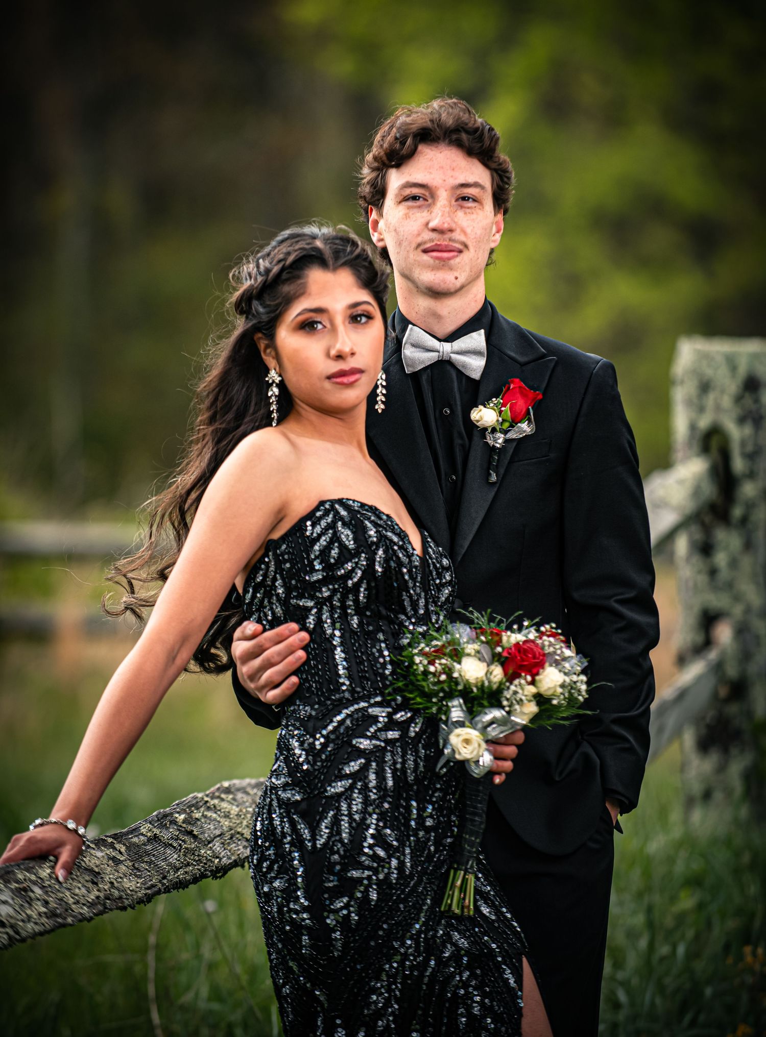 Elegant couple in formal prom attire, woman in black sequin gown, man in tuxedo holding red rose bouquet, outdoor setting.