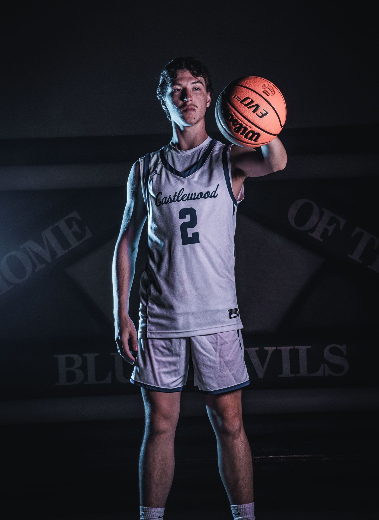 Basketball player in white jersey poses with ball against dark background with team logo visible.
