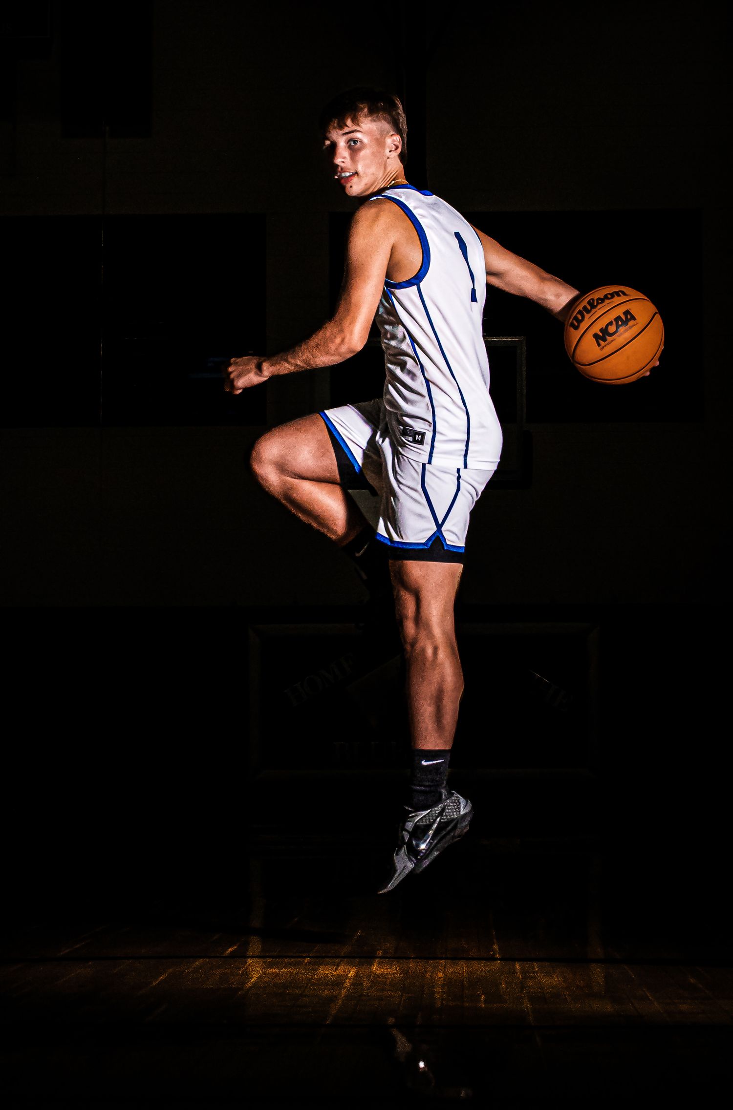 Basketball player in white jersey leaps dramatically while dribbling against dark background.