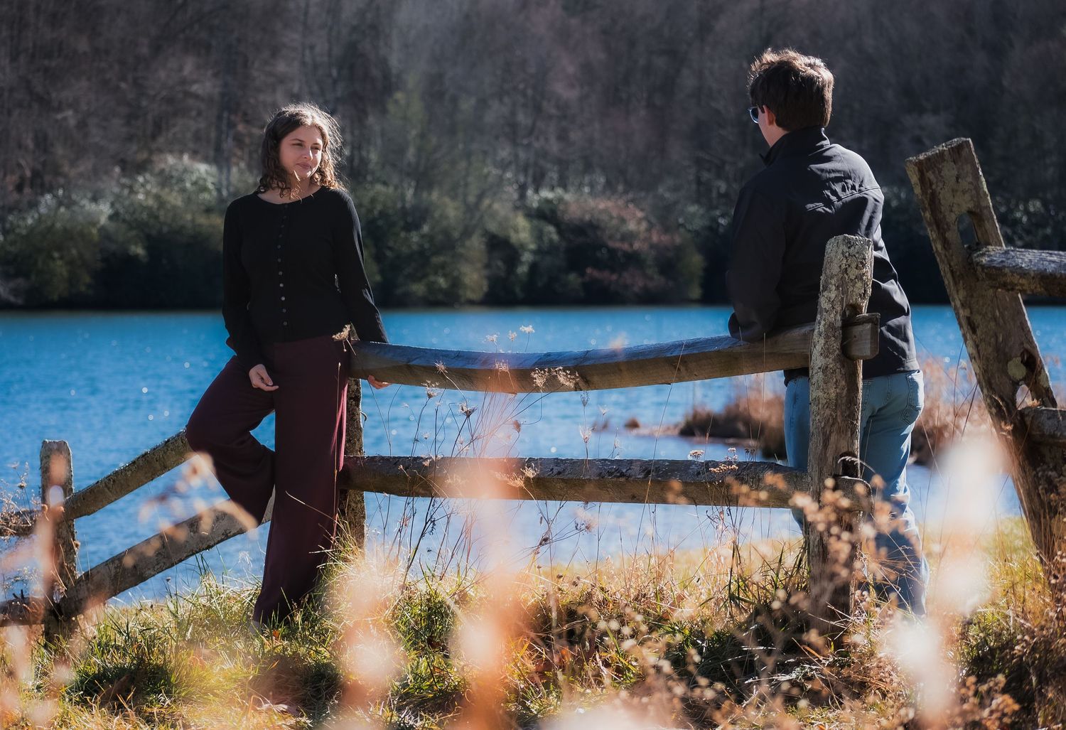 A couple walks together along a scenic lakeside path surrounded by autumn trees and fallen leaves.